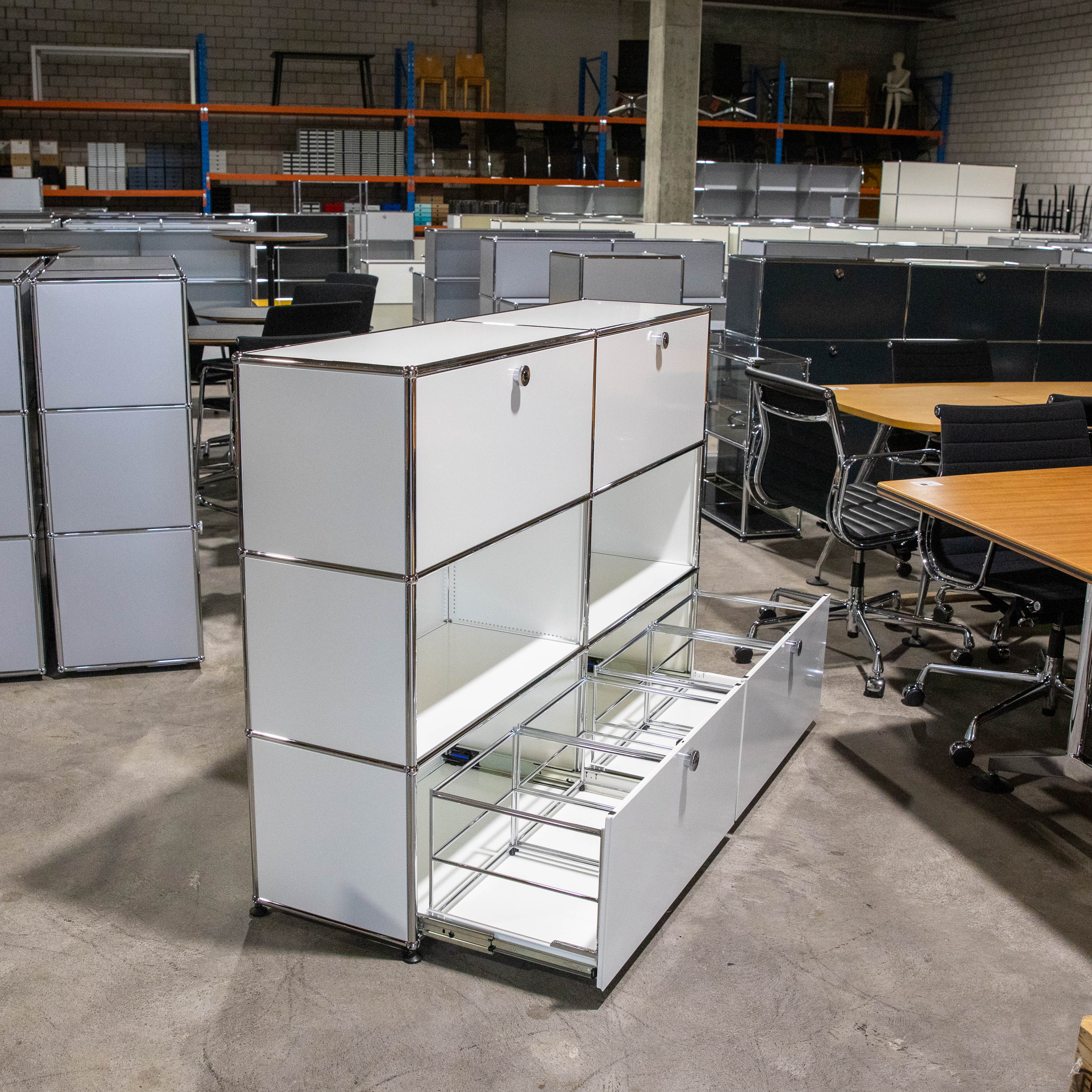 Weisses USM Haller Regal mit 3 Fächern Höhe und 2 Breiten auf Betonboden. Oben geschlossene Klapptüren, unten rechts ein geöffneter Auszug mit integrierter Hängeregistratur. Im Hintergrund weitere USM Möbel.