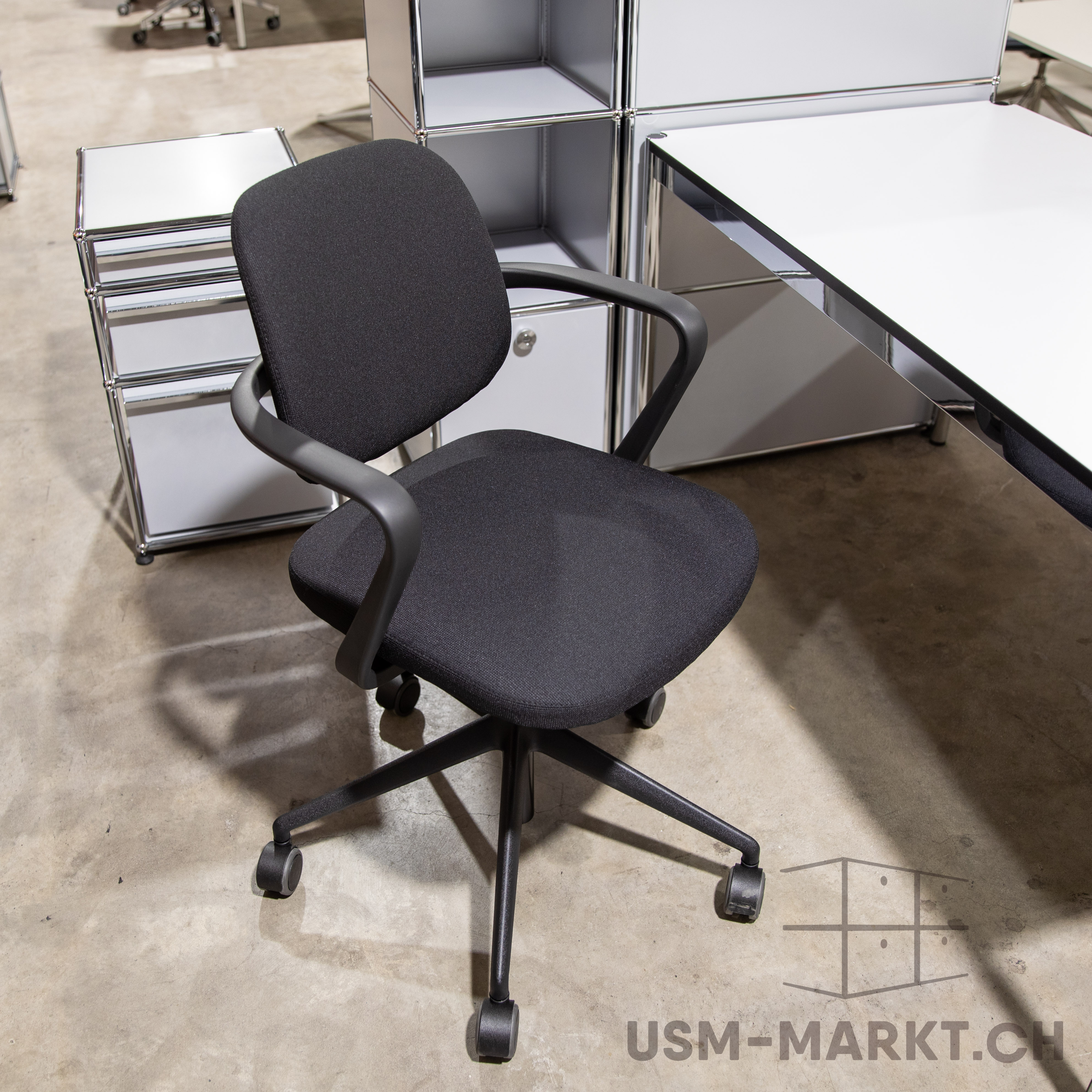 Une chaise de bureau moderne noire, dotée d'accoudoirs et de roulettes, repose sur un sol en béton gris clair. À l'arrière-plan, des meubles USM Haller brillants : un buffet blanc à tiroirs et une étagère à compartiments ouverts.