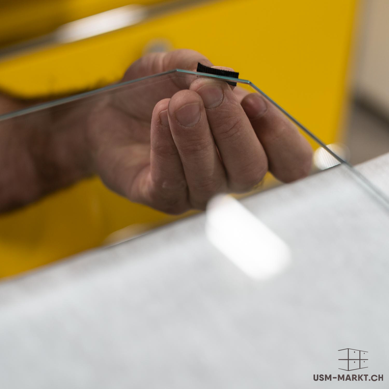 Una mano regge una lastra di vetro trasparente e sottile, con bordo dotato di una piccola protezione nera. Le mani la tengono con precisione, forse per montaggio o controllo qualità. Sullo sfondo sfocato si intravedono superfici gialle.