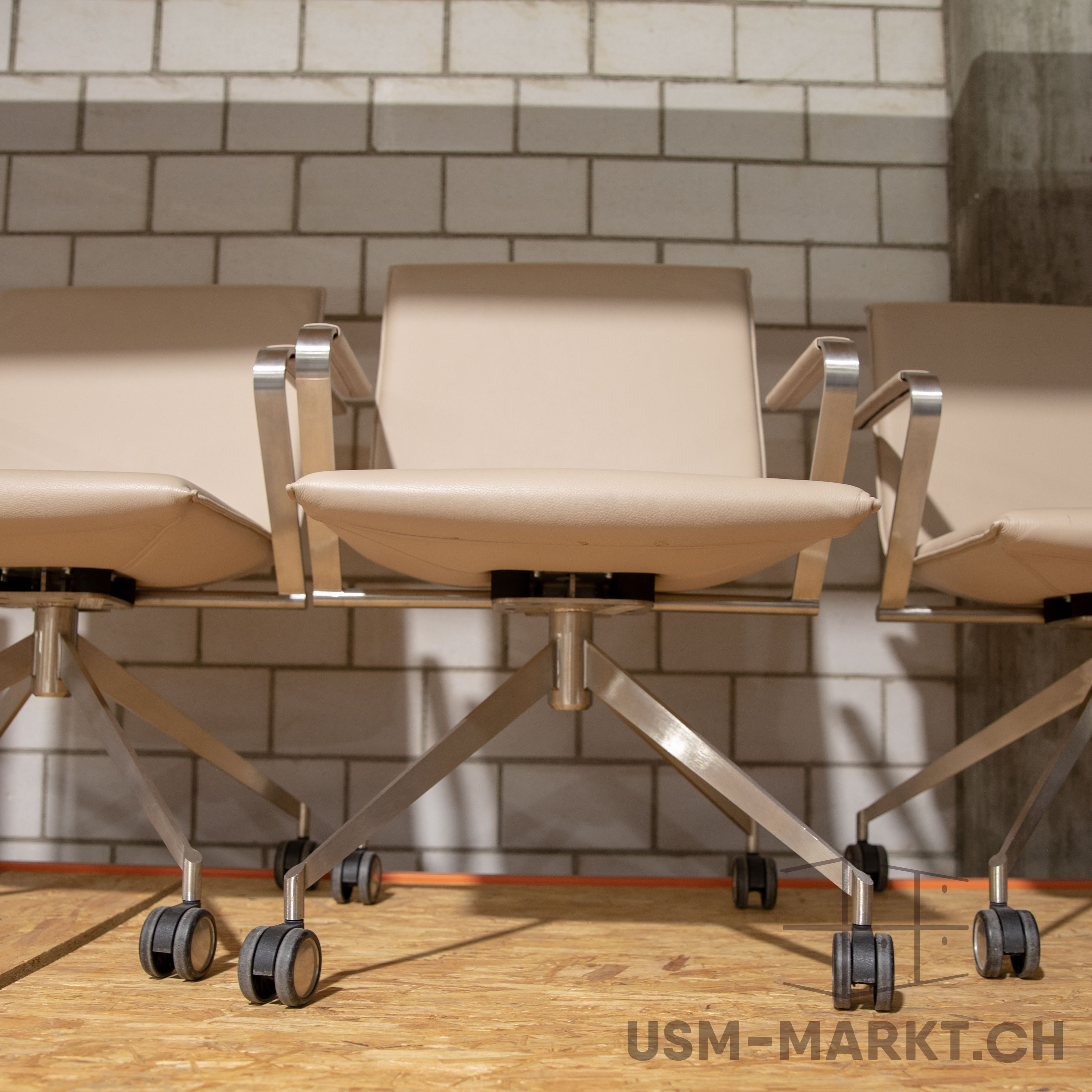 Three modern office chairs in a light beige shade stand side by side on a light brown OSB floor. They have shiny silver metal frames with armrests and black castors. Behind them is a textured white brick wall.