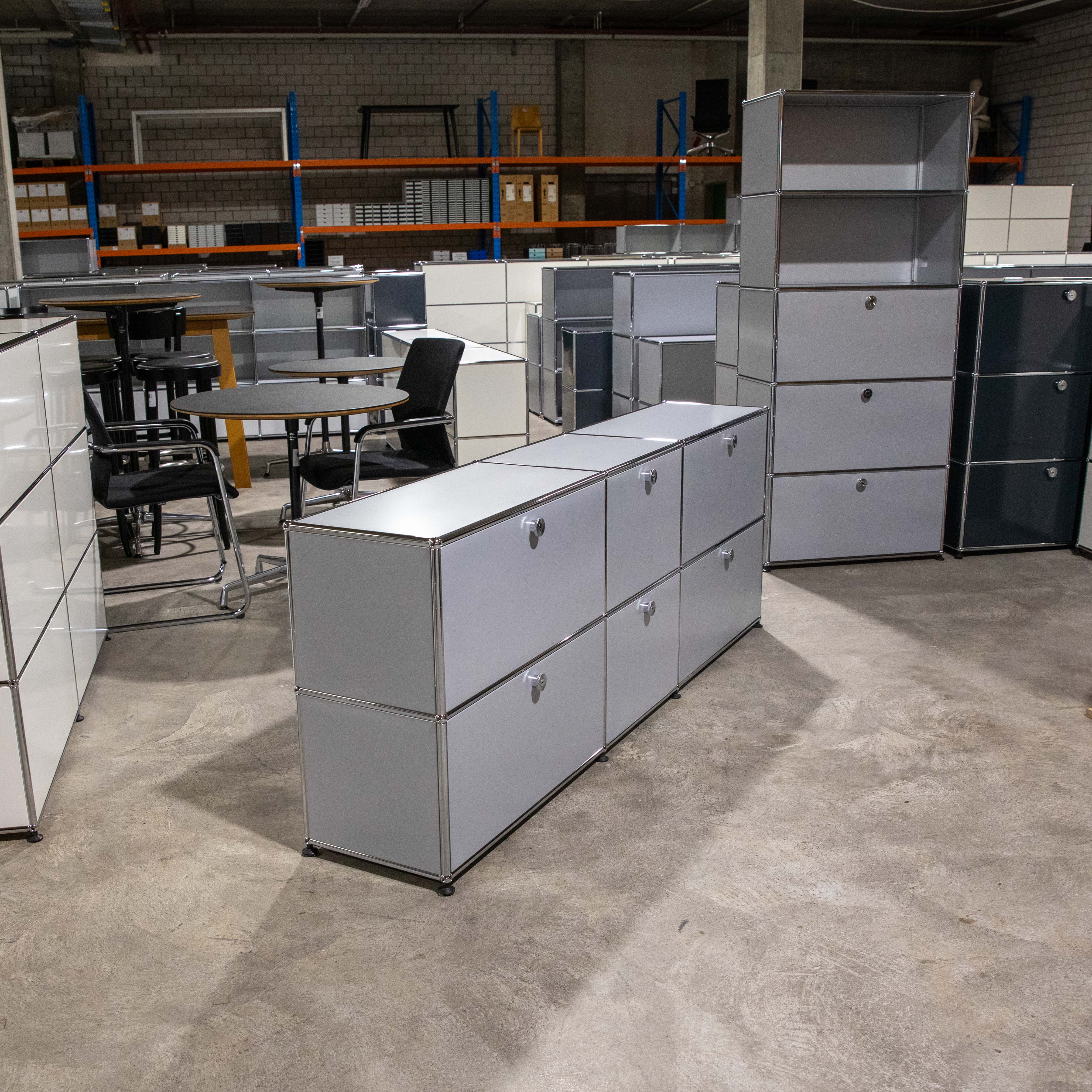 Mattsilbernes USM Haller Sideboard mit zwei Reihen und sechs Modulen auf rauem Betonboden. Drei Klapp- und drei Ausziehtüren mit silbernen Griffen. Im Hintergrund: weitere USM Möbel, Bürostühle, Metallregale im Lagerraum.