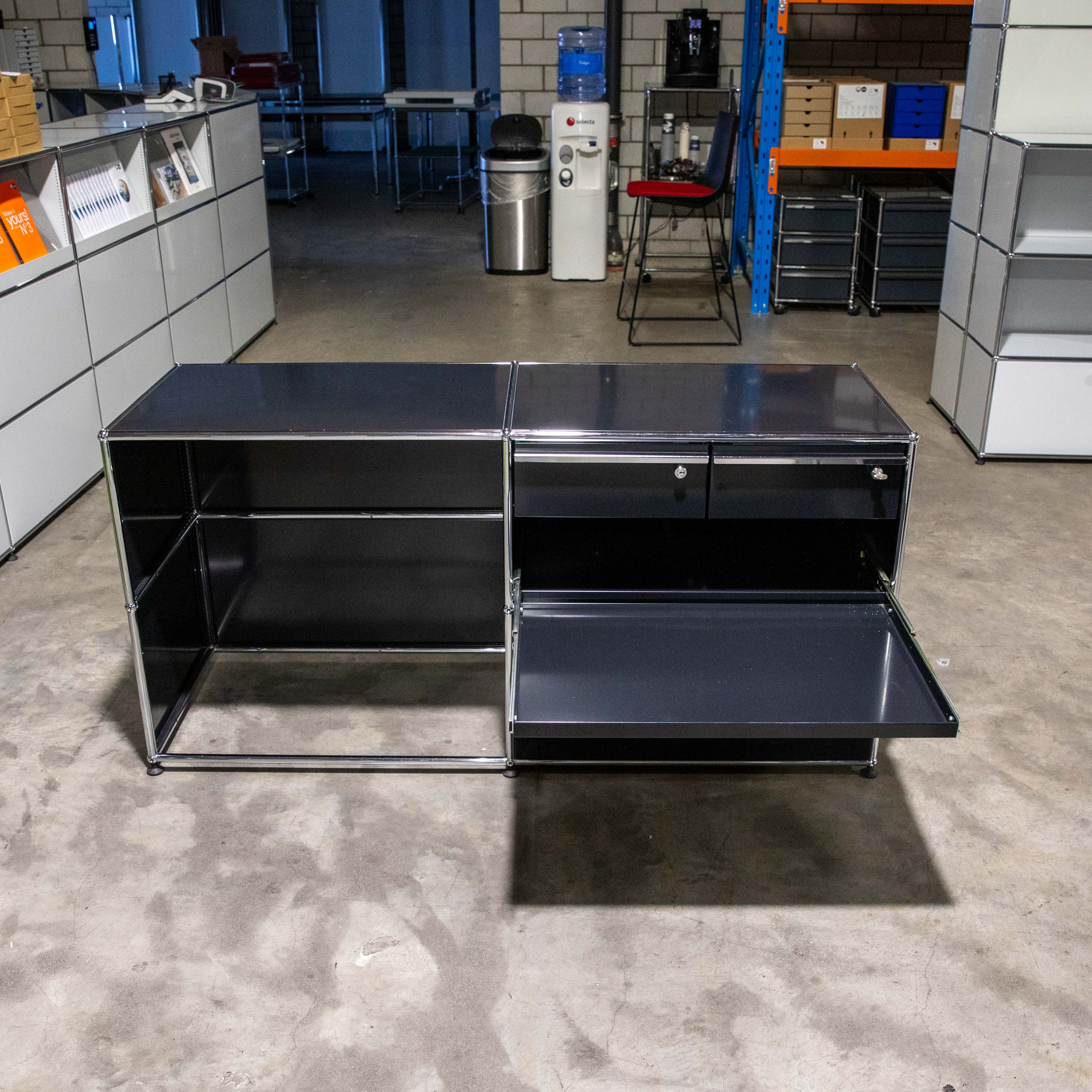 Dunkelgraues, modulares Sideboard mit Chromrahmen auf Betonboden. Zwei Einheiten: links offenes Fach, rechts oben zwei Schubladen und darunter ausgezogene Ablage. Im Hintergrund Büro- und Lagermöbel wie Regale, Wasserspender, Kaffeemaschine.