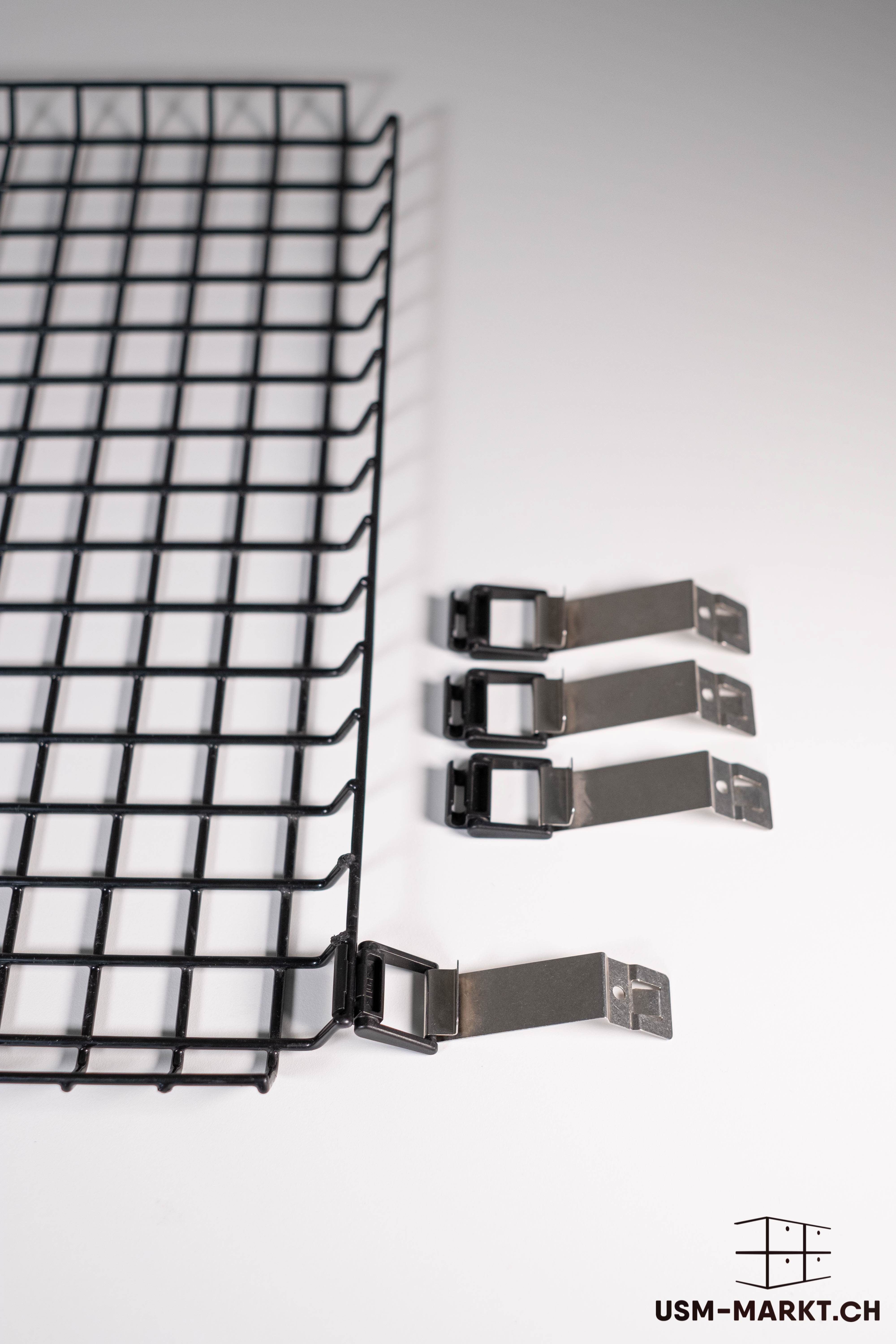 Links ist ein schwarzes Drahtgitterelement mit regelmäßigen rechteckigen Öffnungen zu sehen. Rechts liegen vier silberne Metallklammern mit schwarzen Kunststoffbefestigungen, diagonal angeordnet, zur Montage des Gitters.