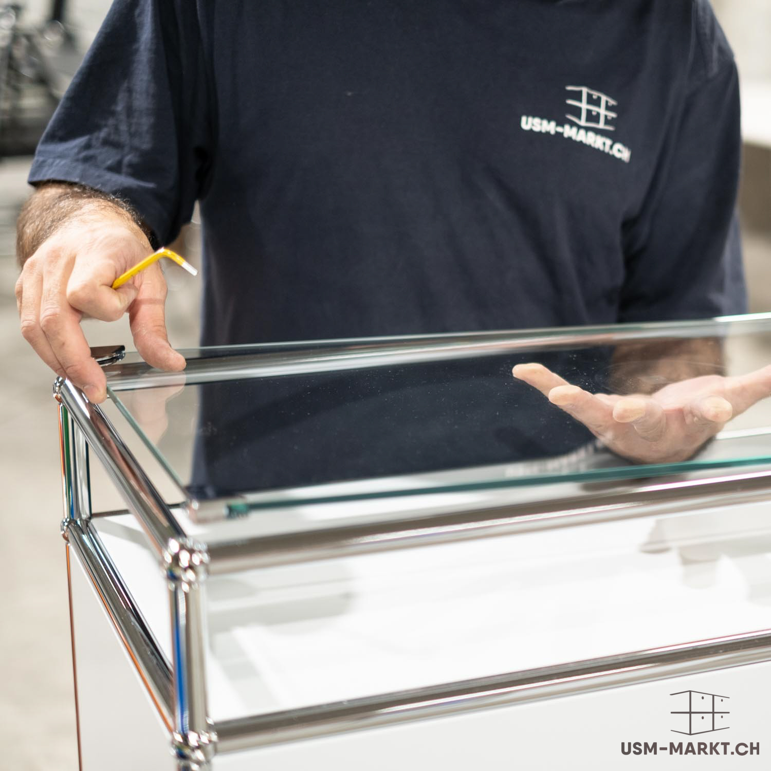 Una persona con una maglietta blu scuro usm-markt sta montando un mobile moderno e modulare. Una mano regge un attrezzo giallo e regola un pannello di vetro, mentre l’altra è sospesa sopra di esso. Tubi cromati e pannelli di vetro bianchi e trasparenti.