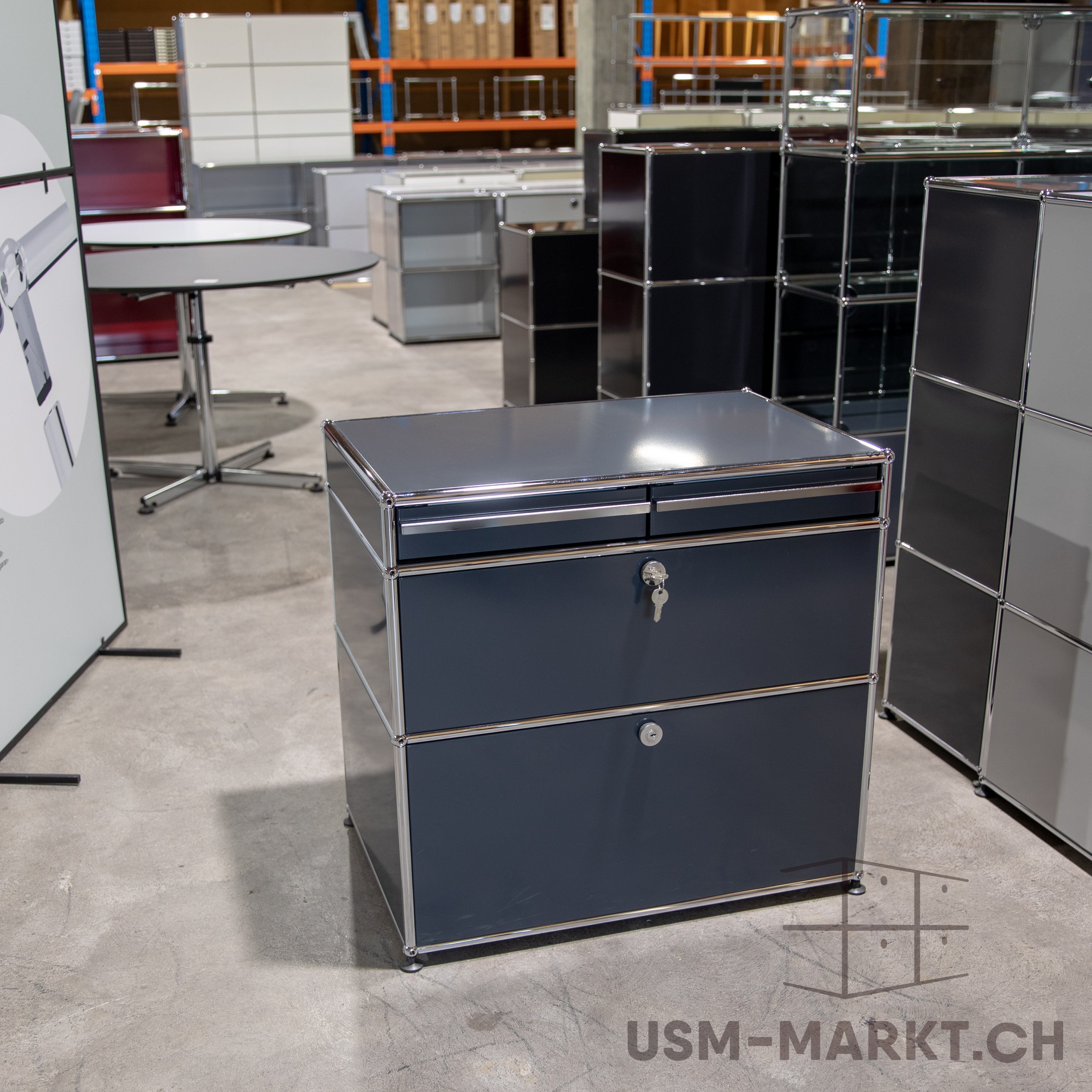 Dunkelgrauer modularer Aktenschrank mit zwei Schubladen und polierten Chromrahmen auf hellgrauem Betonboden. In der oberen Schublade steckt ein kleiner Schlüssel. Unscharf im Hintergrund: weitere Möbel, Tische und Regale.
