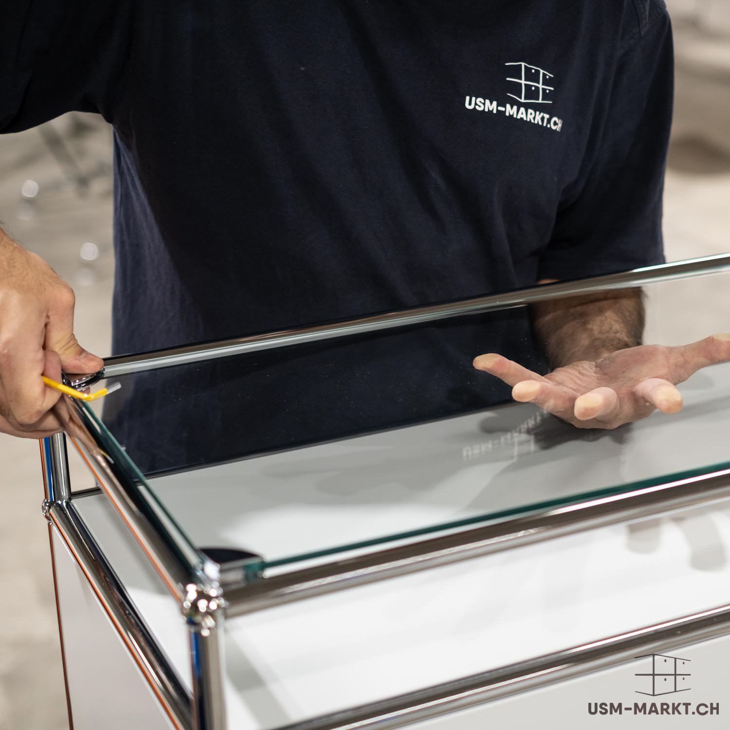 Una persona con maglietta blu scuro con logo usm-markt.ch monta un pannello di vetro trasparente su un mobile USM Haller bianco con struttura cromata. Con la sinistra lo allinea con un attrezzo giallo, con la destra lo sostiene.