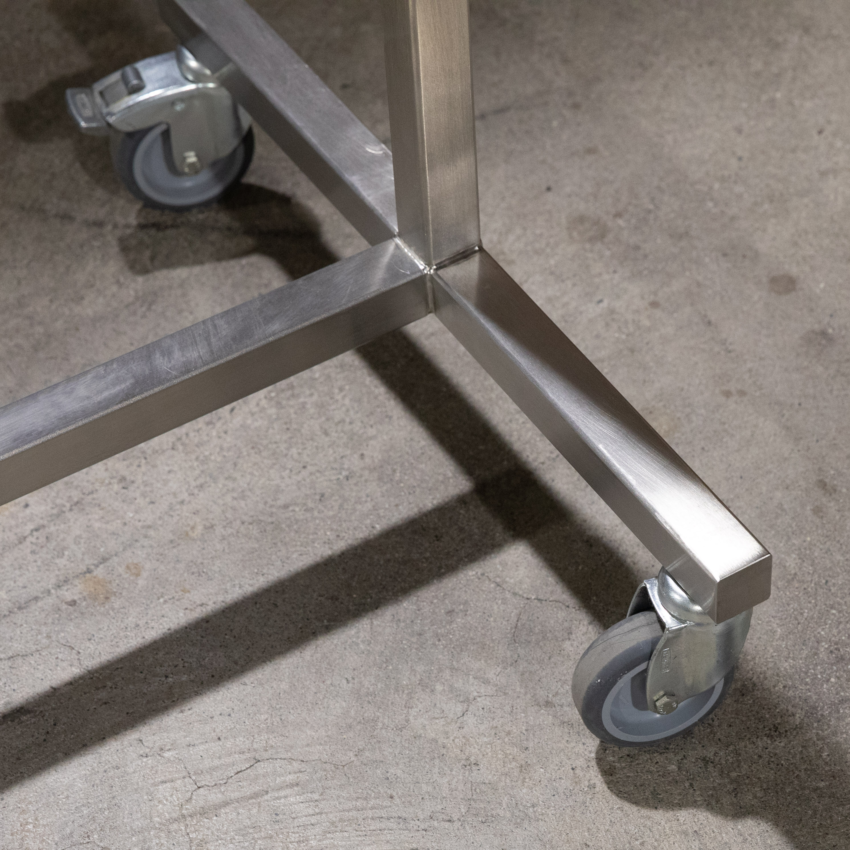 Nahaufnahme des unteren Bereichs eines Kleiderständers aus gebürstetem Chromstahl auf Rollen. Verschweißte Metallstreben bilden eine stabile Basis. Eine graue, arretierbare Rolle steht auf grauem Betonboden. Leichte Gebrauchsspuren.
