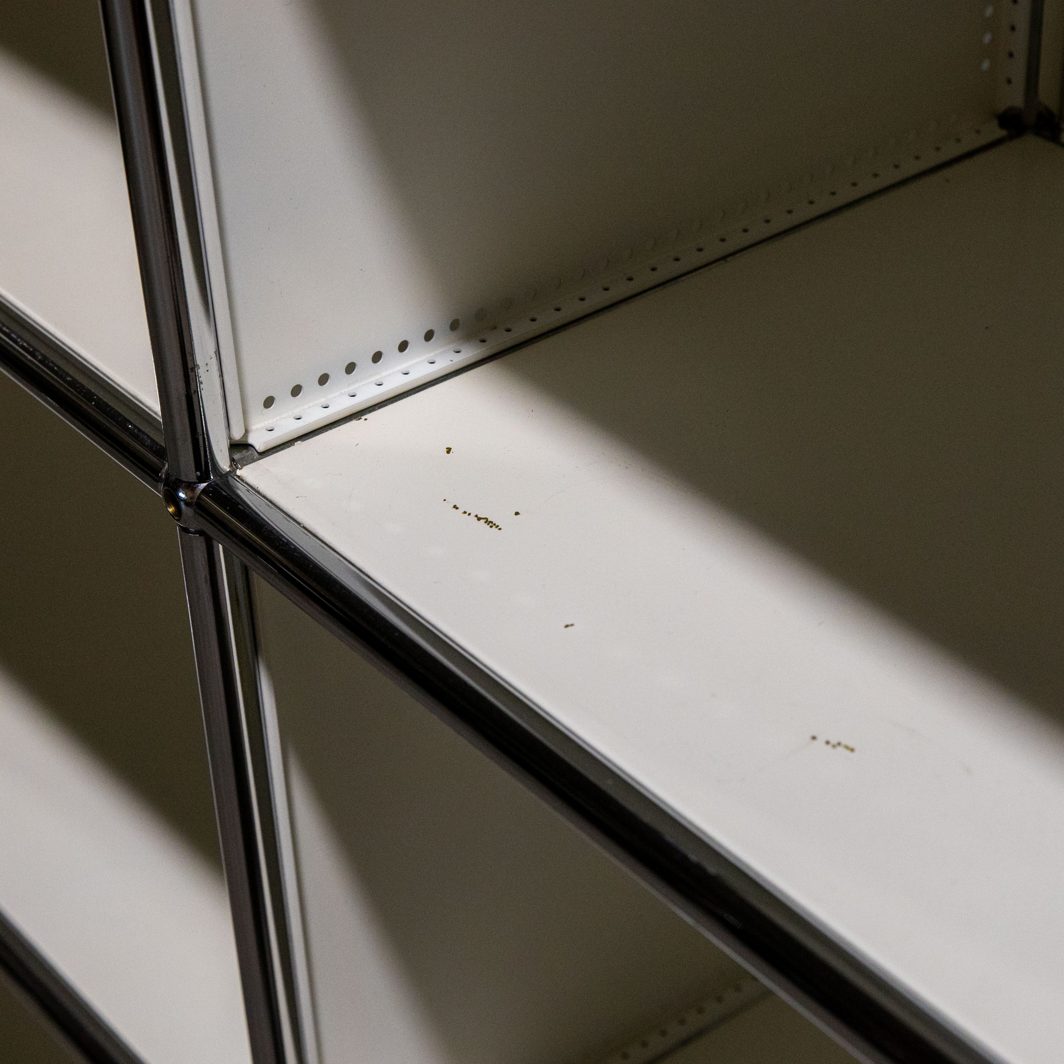 Detailansicht eines modularen Regalsystems mit glänzenden, verchromten Verbindungsrohren und matten, weißen Metallplatten. Feiner Staub und kleine Flecken sind sichtbar. Perforierte Hinterkanten; diagonal verlaufender Schatten.