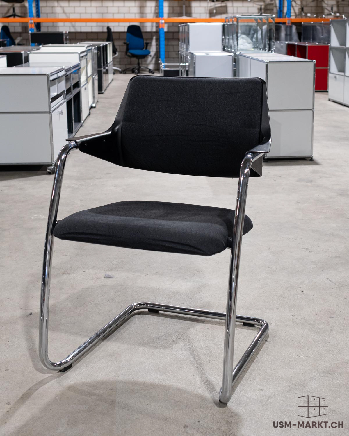 Ein schwarzer Freischwinger mit glänzendem Chromgestell steht auf grauem Betonboden. Gepolsterte schwarze Sitzfläche und Rückenlehne. Im Hintergrund Reihen weißer modularer Lagerschränke und ein blauer Bürostuhl.