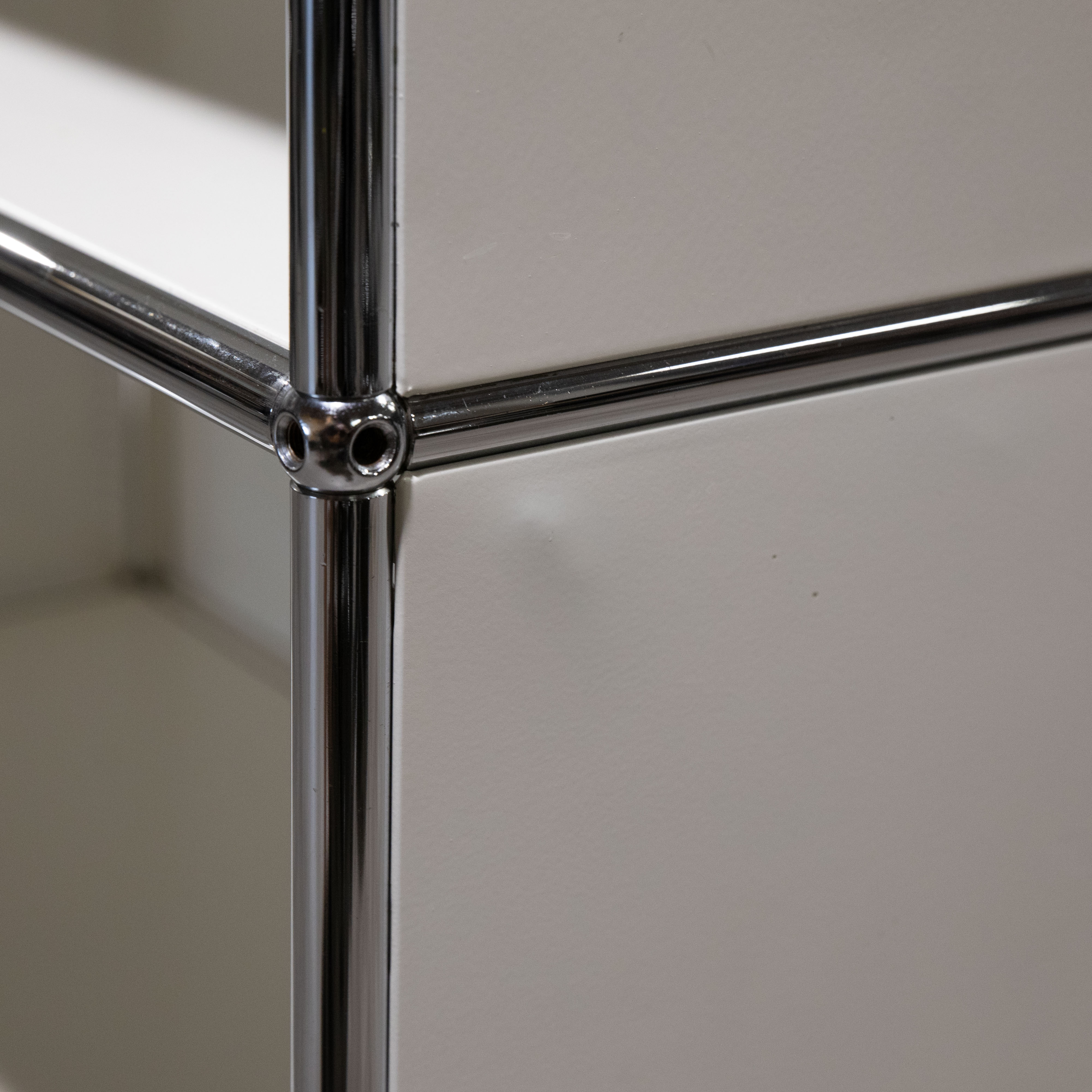 Nahaufnahme einer modularen Regalstruktur aus glänzenden, verchromten Stahlrohren mit kugelförmigen Verbindern. Weiße, glatte Paneele bilden die Fächer. Ecke der Einheit im Fokus, mit weichen Schatten und Licht.