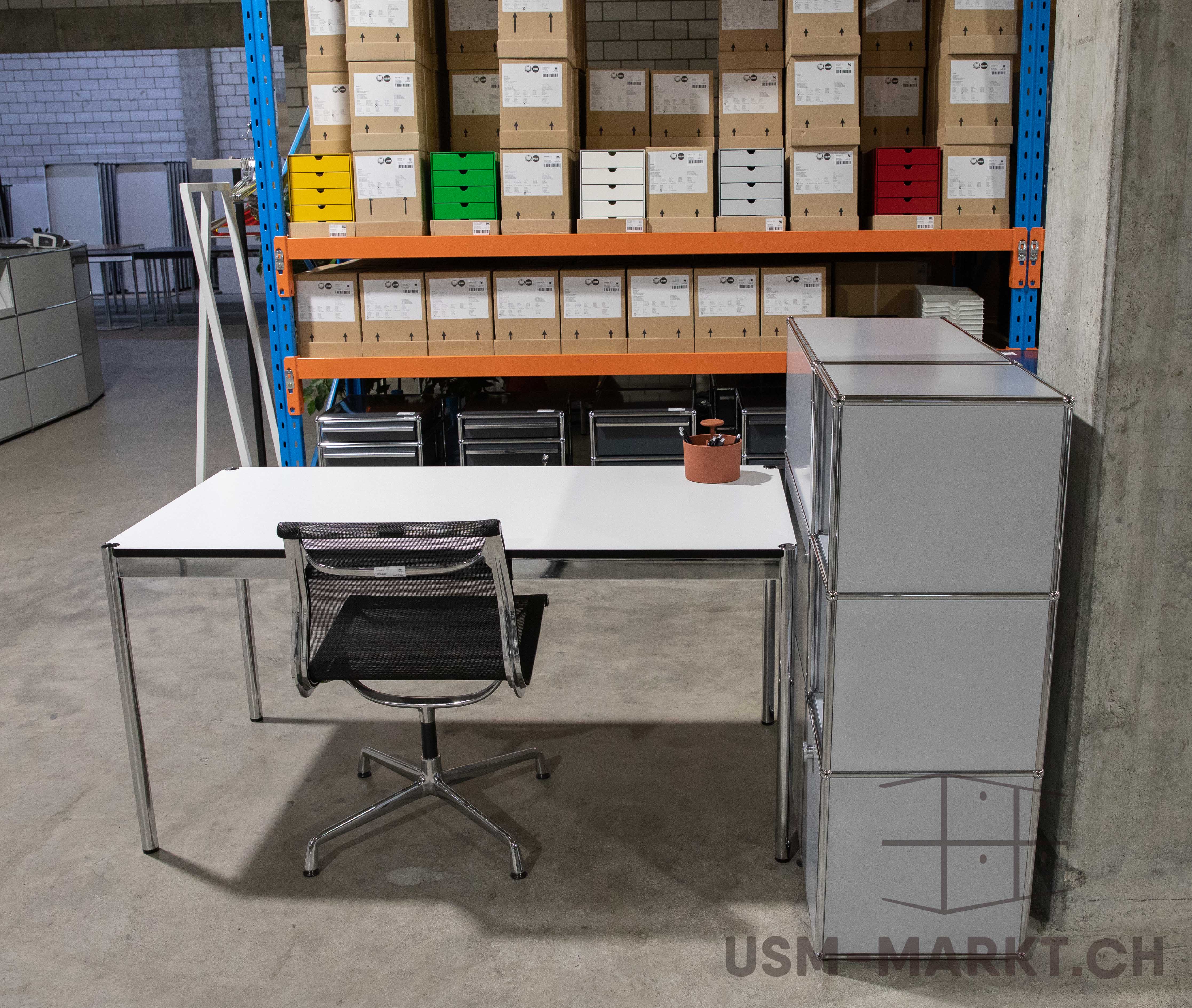 Moderner weißer Schreibtisch und schwarzer Bürostuhl in einem Lagerraum. Hinten ein Regal mit braunen Kartons und bunten Schubladen in Gelb, Grün, Weiß und Rot. Rechts ein hoher grauer Modularschrank. Stiftehalter auf dem Tisch.