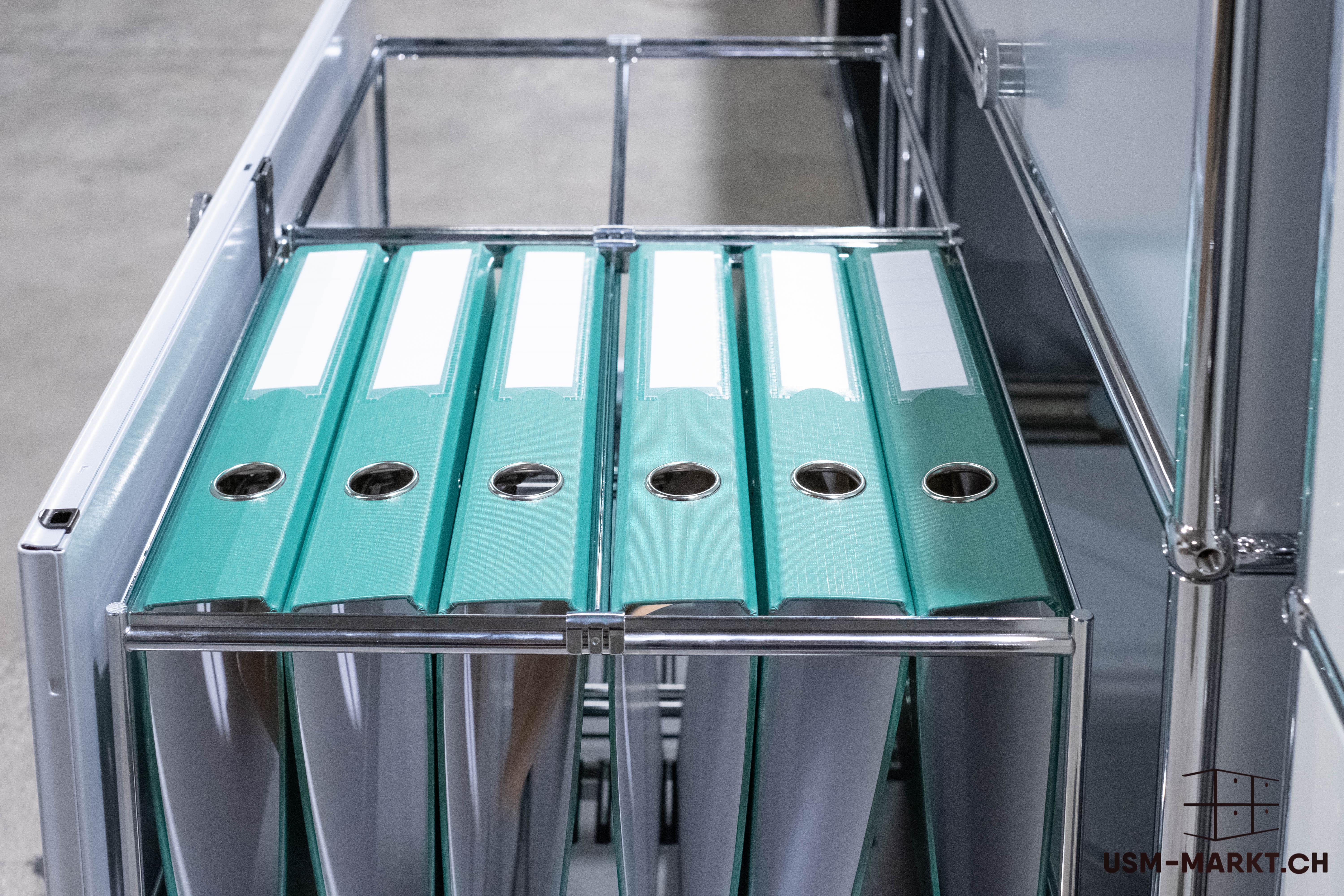 Six mint-green file folders with white labels stand close together on a pull-out shelf in a modern office unit. Viewed from above, the finger holes and labels are clearly visible.
