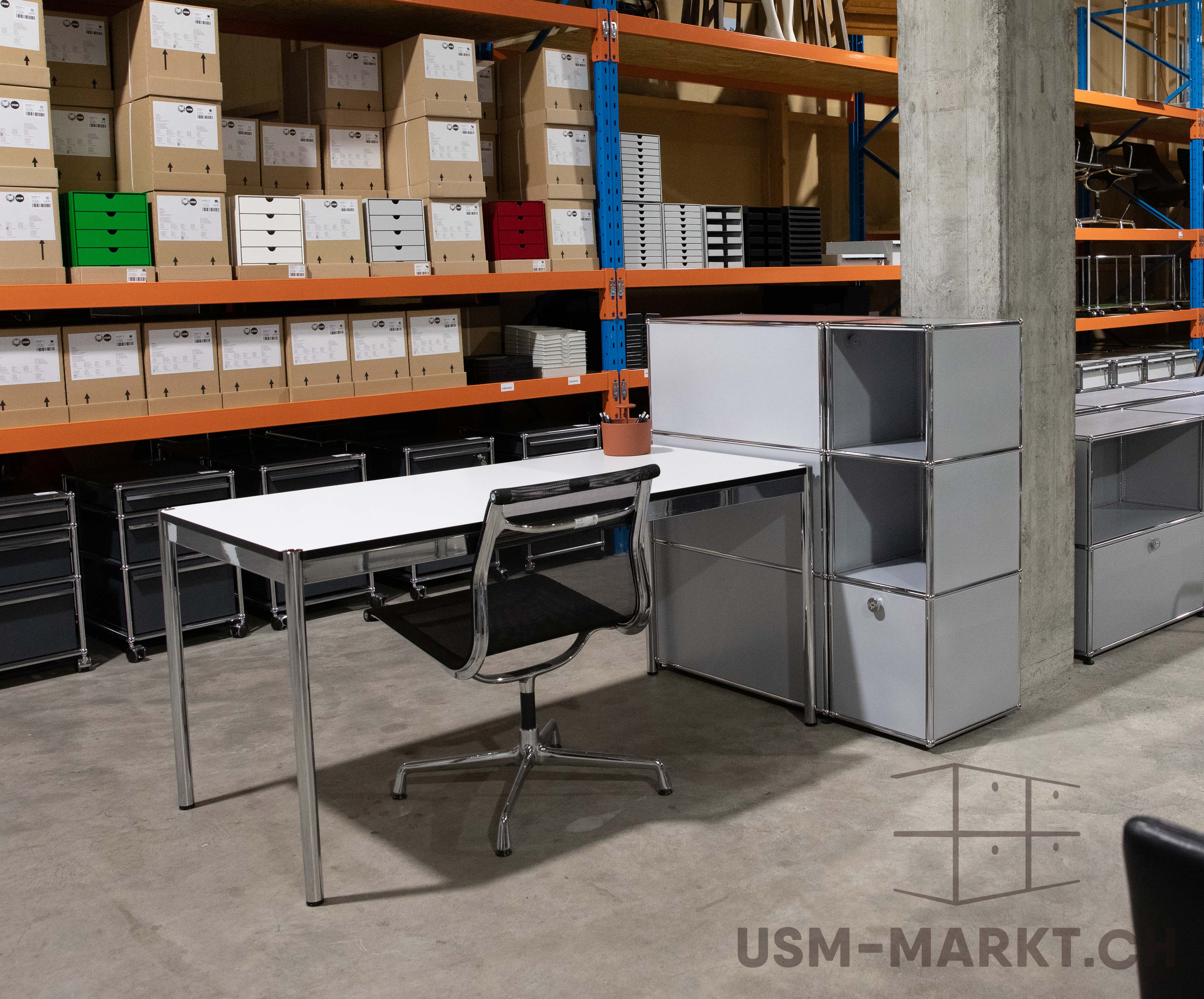 Modernes Büro-Set mit weißem Schreibtisch und schwarzem Netzstuhl im Vordergrund eines Warenlagers. Im Hintergrund hohe orange Regale mit braunen Kartons und bunten Ablageboxen. Rechts modulare hellgraue Büroschränke.