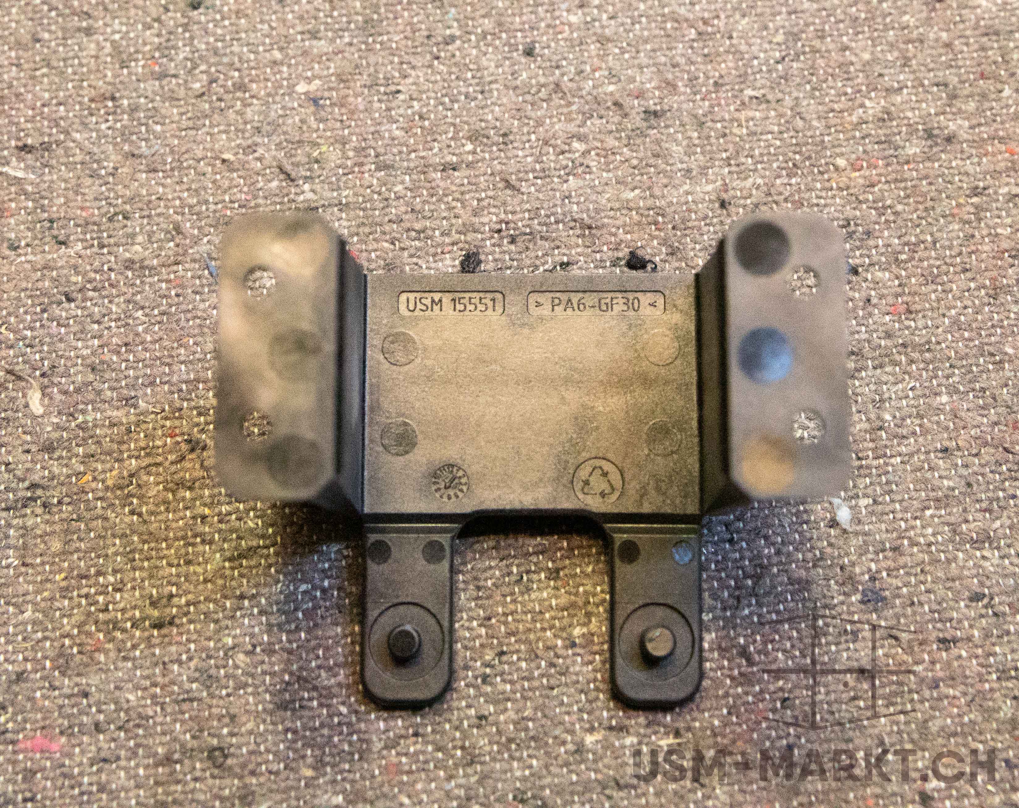 Dark grey, angled plastic component with a flat central plate, embossed with ‘USM 15551’, ‘PA6-GF30’ and the recycling symbol. Flanges curved upwards on both sides with round recesses; two legs projecting downwards with round dots.