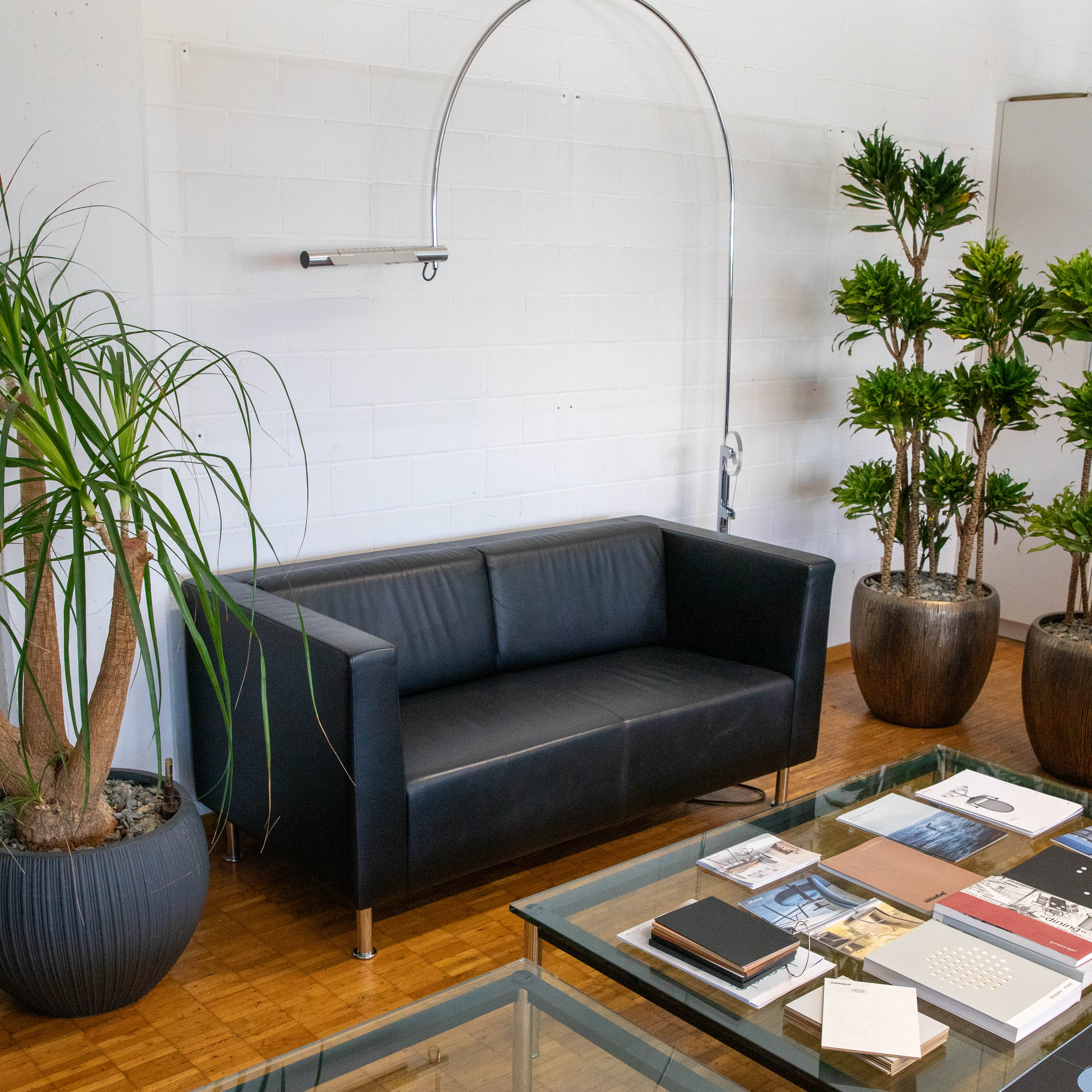 Baltensweiler ’s Halo arc lamp, with its curved silver metal arm, towers over a black leather sofa. In front of it stands a glass table with books on it. Tall green plants in dark pots frame the scene, set against a white brick wall and wooden floor.