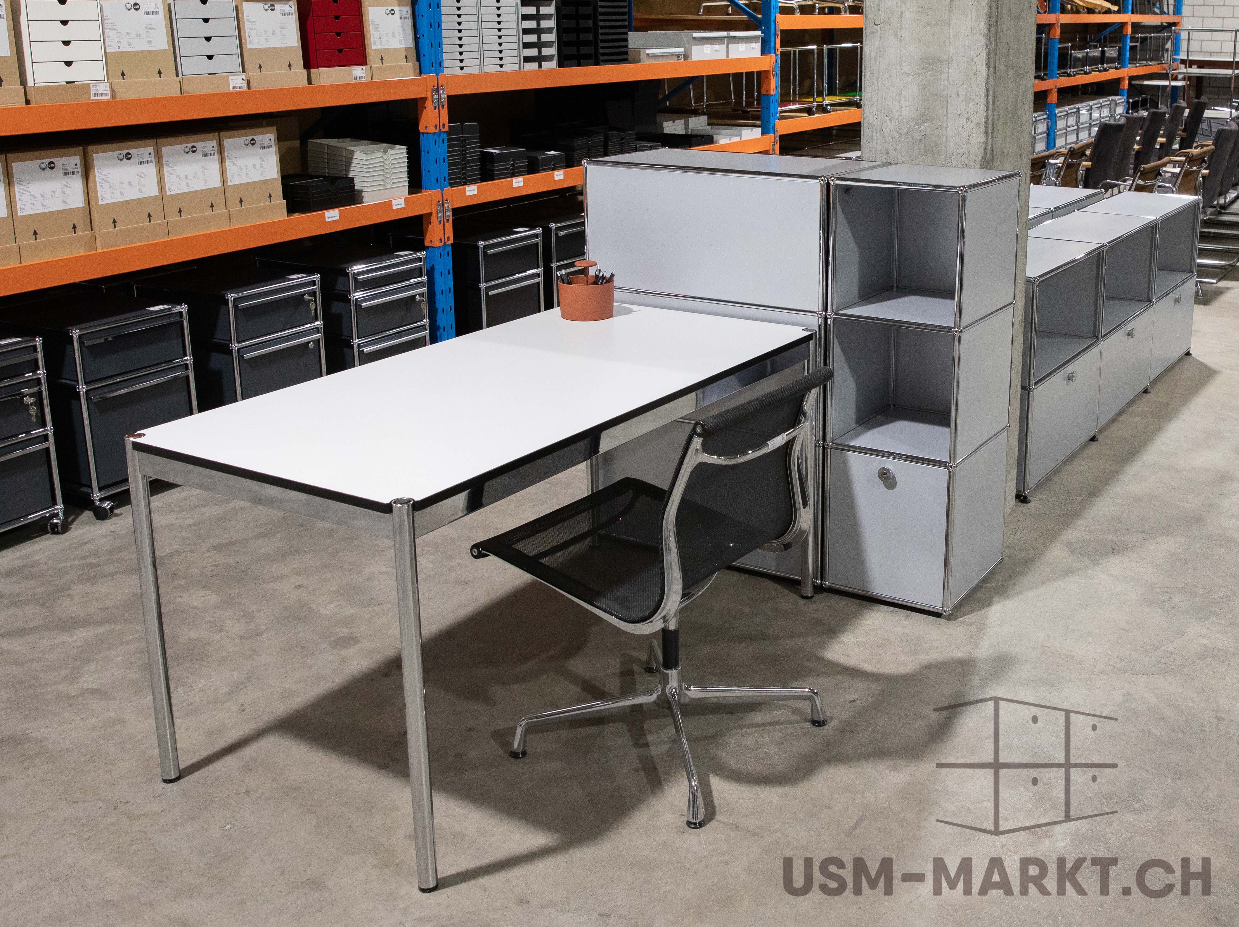 Arbeitsplatz in einem Lager mit weissem USM Haller Tisch, schwarzem Bürostuhl und modularen silbernen USM Haller Sideboards. Im Hintergrund hohe orangefarbene Regale mit Kartons und Möbelteilen, industrielles Umfeld.