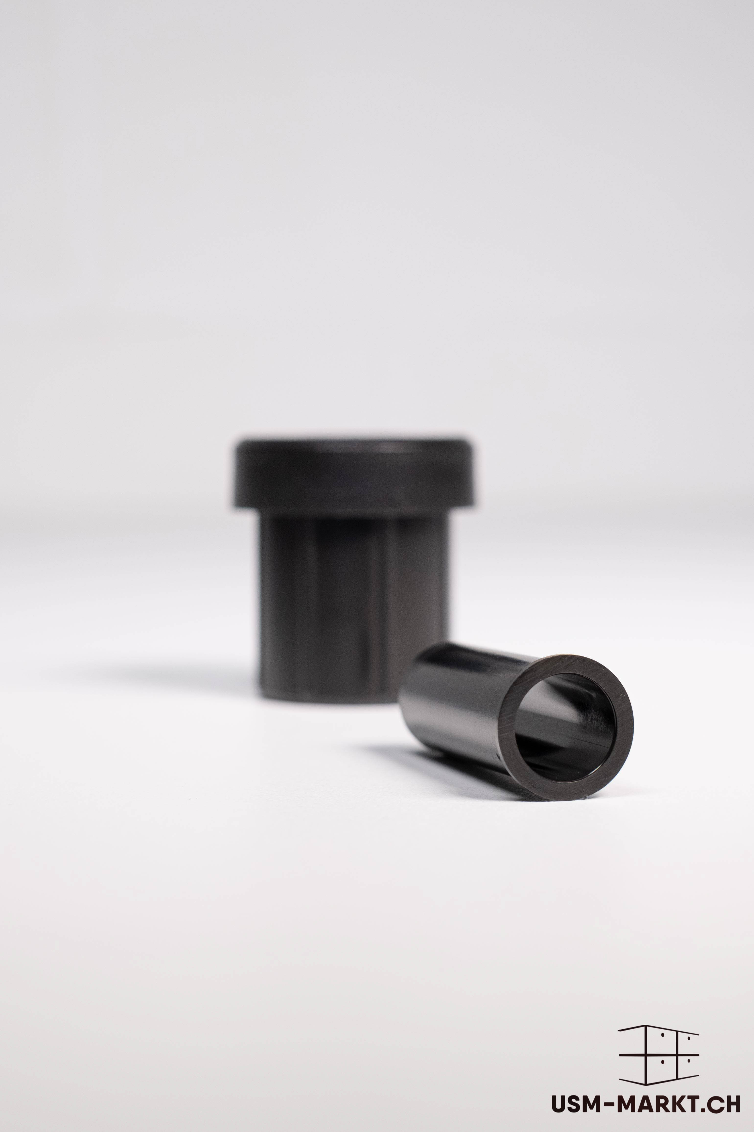 Two black, cylindrical connectors are shown against a light, plain background. In the foreground, a hollow sleeve lies horizontally with a visible round opening; behind it, a wider socket stands vertically, slightly out of focus.