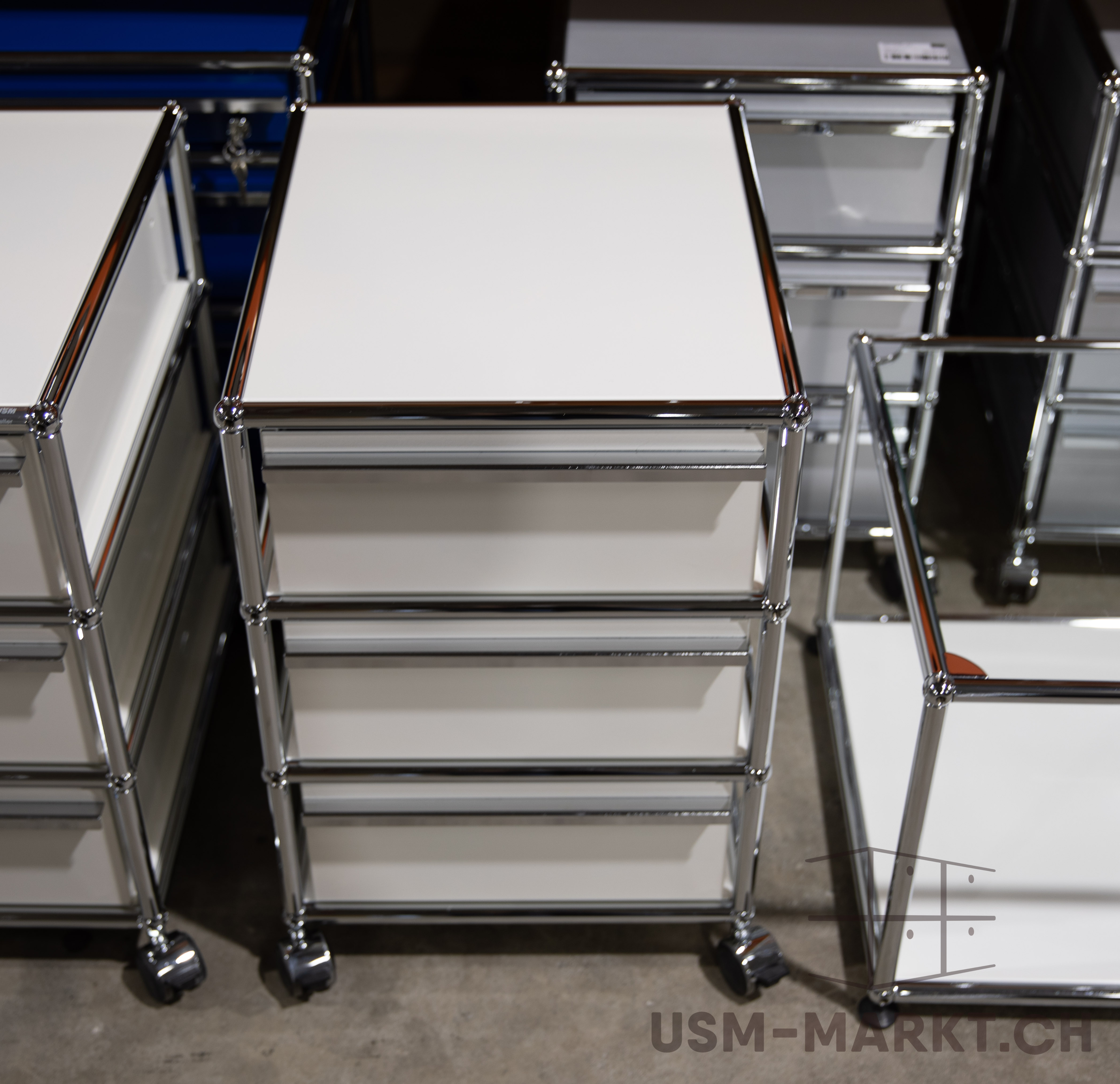 Un moderno carrello portadocumenti bianco con quattro cassetti e struttura cromata è su un pavimento in cemento grigio. Sullo sfondo si vedono mobili simili, bianchi e cromati, come in un magazzino o showroom.