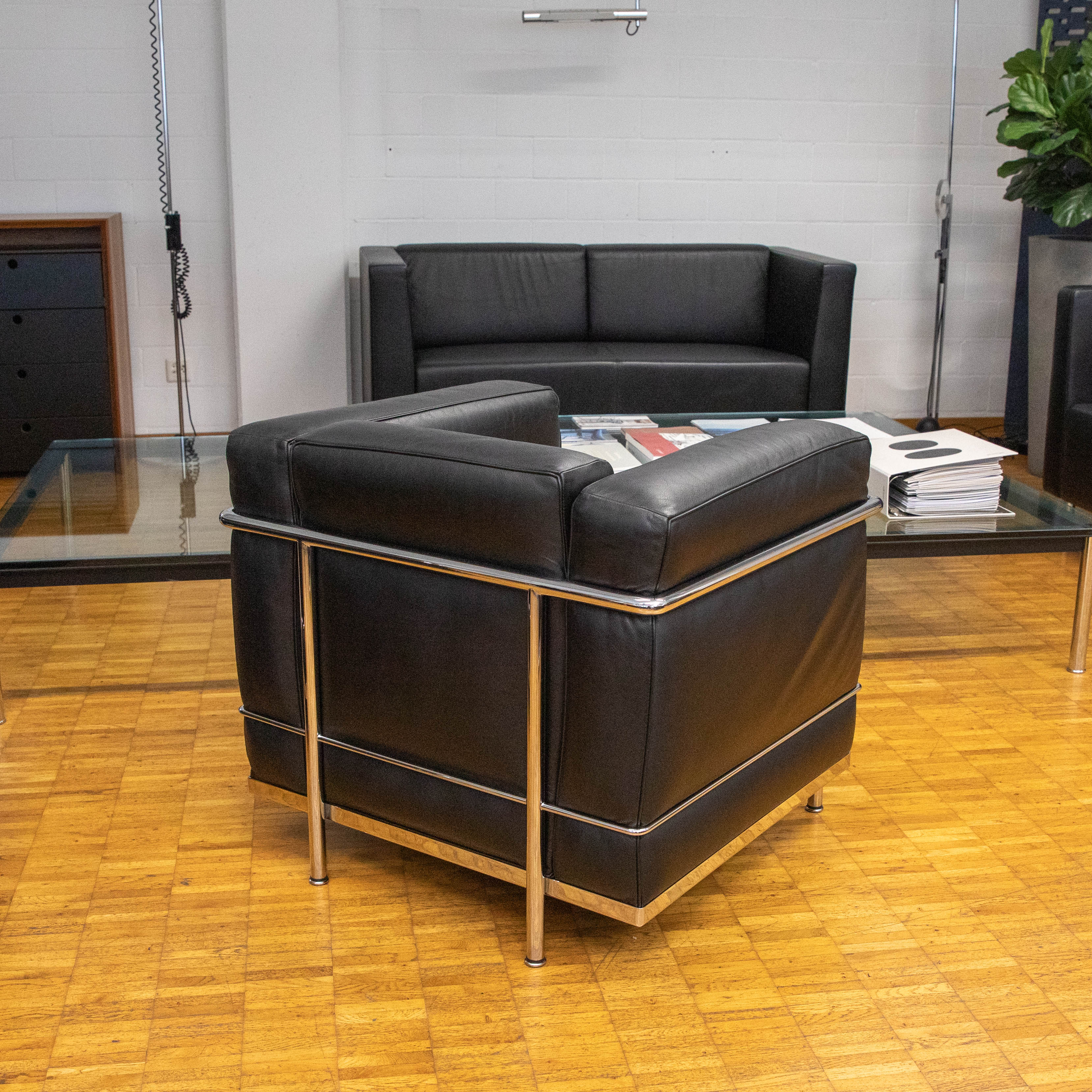 A black Le Corbusier LC2 leather armchair from Cassina stands in the foreground on a parquet floor. The armchair has a polished chrome frame. In the background, a black leather sofa and a glass table with magazines can be seen.