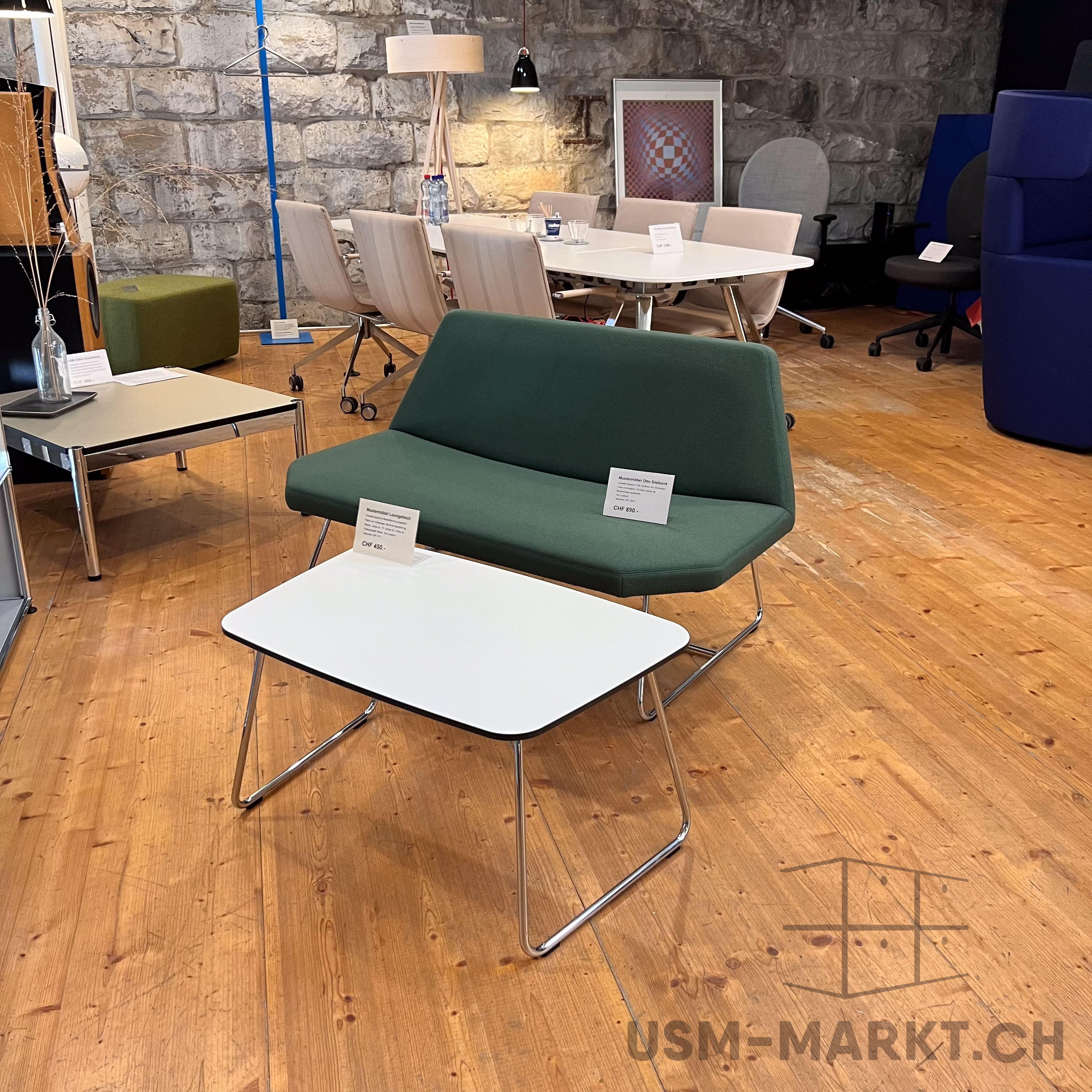 Grüne Polsterbank mit Metallgestell auf Holzboden, davor ein kleiner weißer Couchtisch. Im Hintergrund eine rustikale Steinwand, davor ein großer Konferenztisch mit hellen Stühlen. Büromöbel und moderne Beleuchtung ergänzen den Raum.