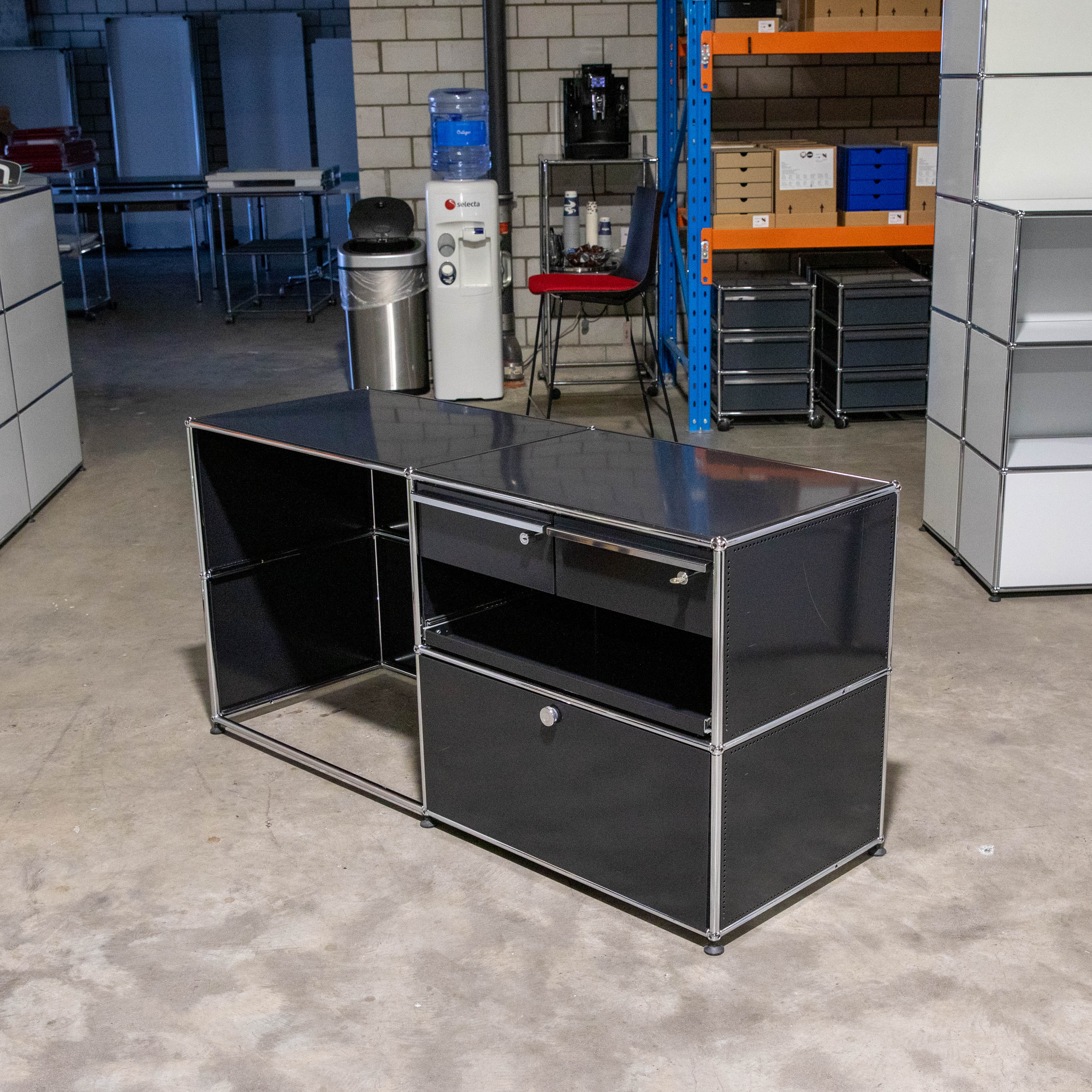 Schwarzer modularer Schreibtisch/Kommode mit Chromrahmen. Links offene Fächer, zwei Schubladen, rechts abschließbarer Schrank. Betonboden; im Hintergrund Wasserspender, Kaffeemaschine und Regale.