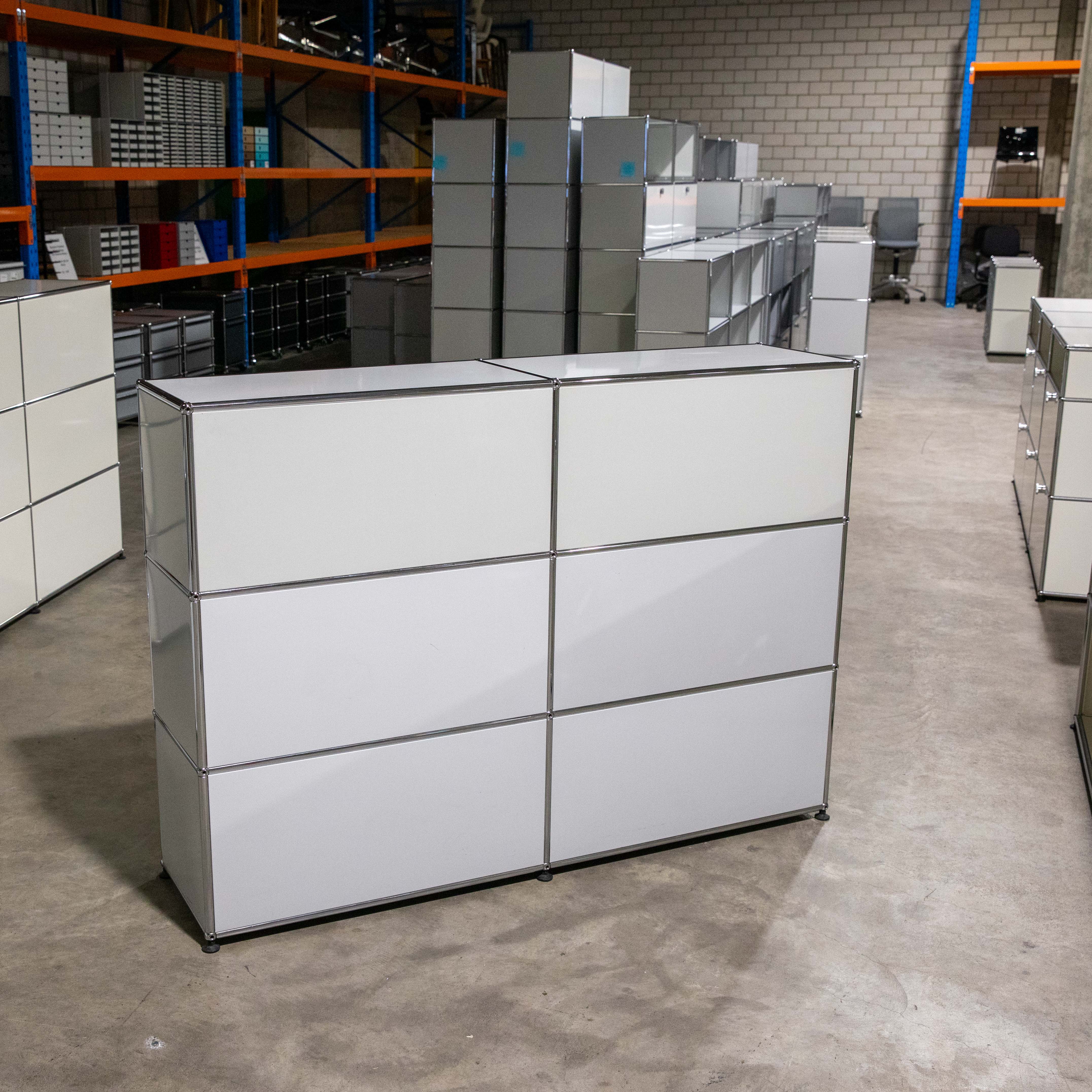 A light grey USM Haller sideboard with six compartments (3 rows, 2 columns) on a light grey concrete floor in a warehouse. Smooth metal fronts, framed by polished chrome tubes. Modular furniture and orange storage shelving in the background.