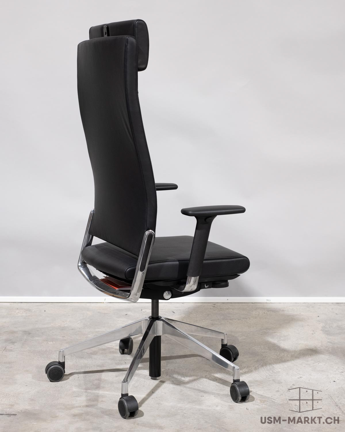 Une élégante chaise de bureau haute en cuir noir trône sur un sol en béton gris clair, devant un mur sobre. Forme ergonomique avec appui-tête intégré, accoudoirs confortables et piètement chromé à cinq roulettes.
