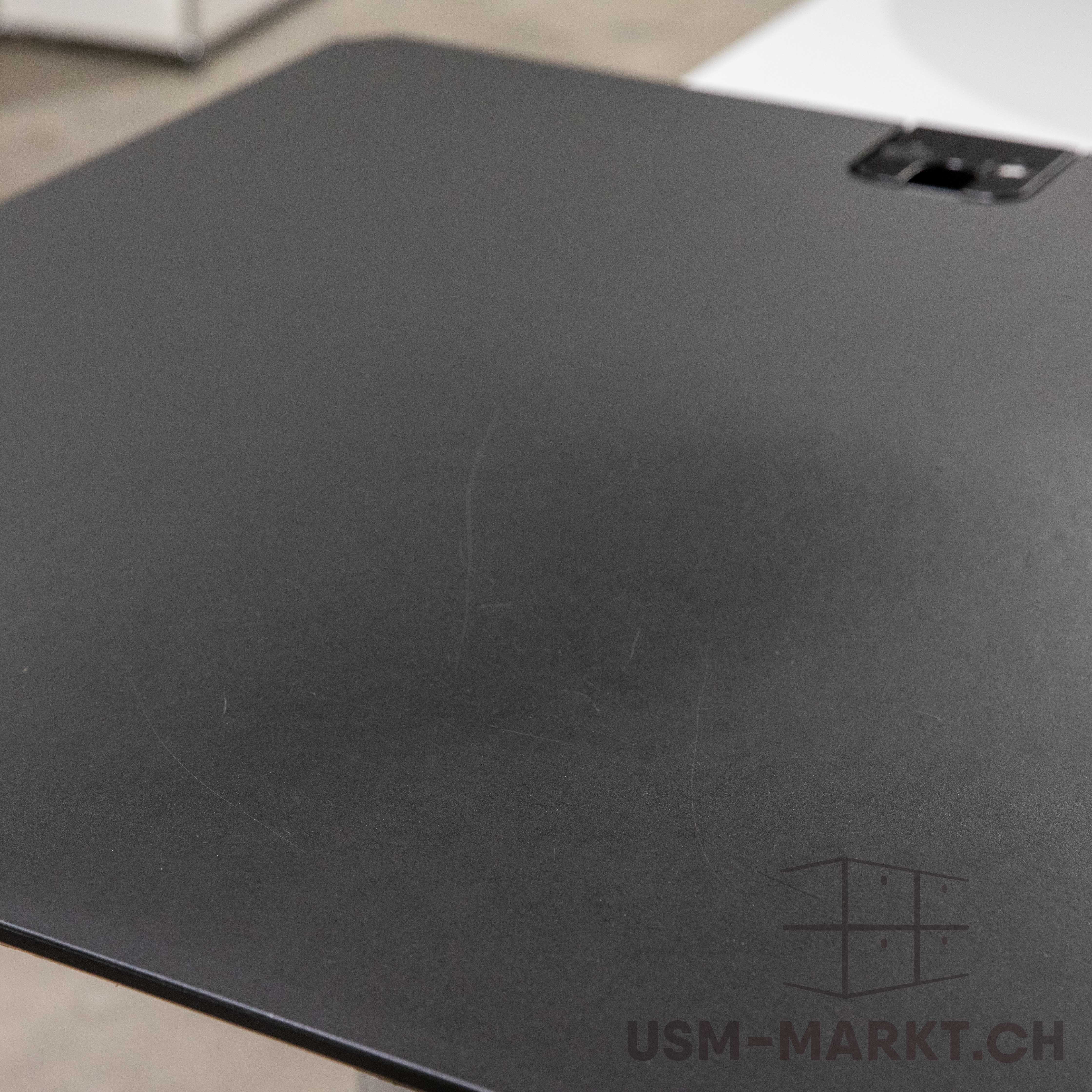 Nahaufnahme der schwarzen Linoleum-Tischplatte eines gebrauchten USM Kitos E Plus. Die Oberfläche weist mehrere feine Kratzer und Gebrauchsspuren auf. Im oberen rechten Bereich ist eine offene Klappe zu sehen, die einen integrierten Kabelkanal freigibt.