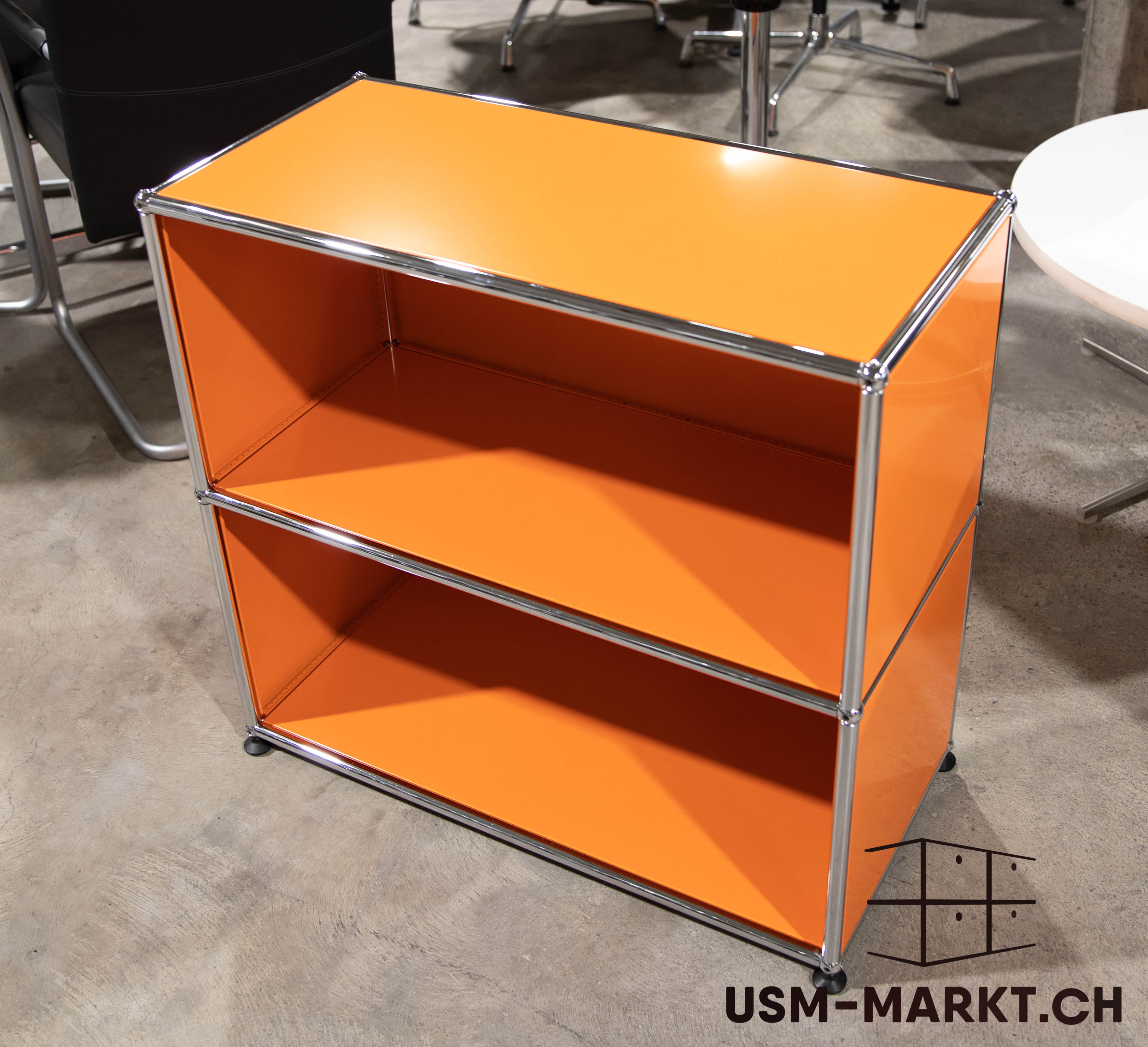 Ein leuchtend orangefarbenes, modulares Regal mit zwei offenen Fächern und glänzenden Chromrahmen steht auf hellgrauem Betonboden. Im Hintergrund sind unscharf ein schwarzer Bürostuhl und ein runder weißer Tisch zu erkennen.
