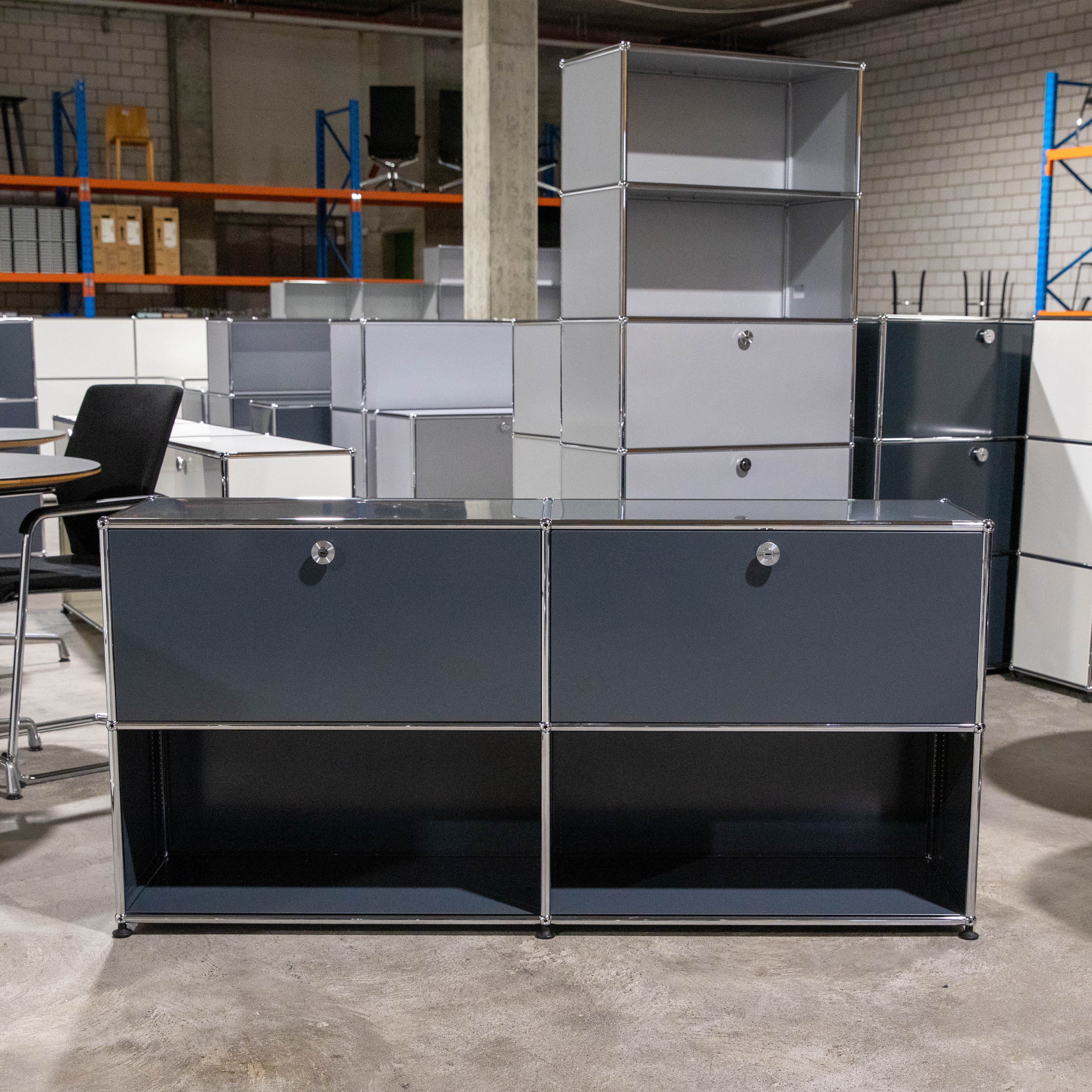 Anthrazitfarbenes USM Haller Sideboard mit modularem Chromstahlrohrsystem, zwei Felder breit und zwei Fächer hoch. Oben zwei geschlossene Klapptüren mit silbernen Griffen, unten zwei offene Ablagefächer. Auf Betonboden.