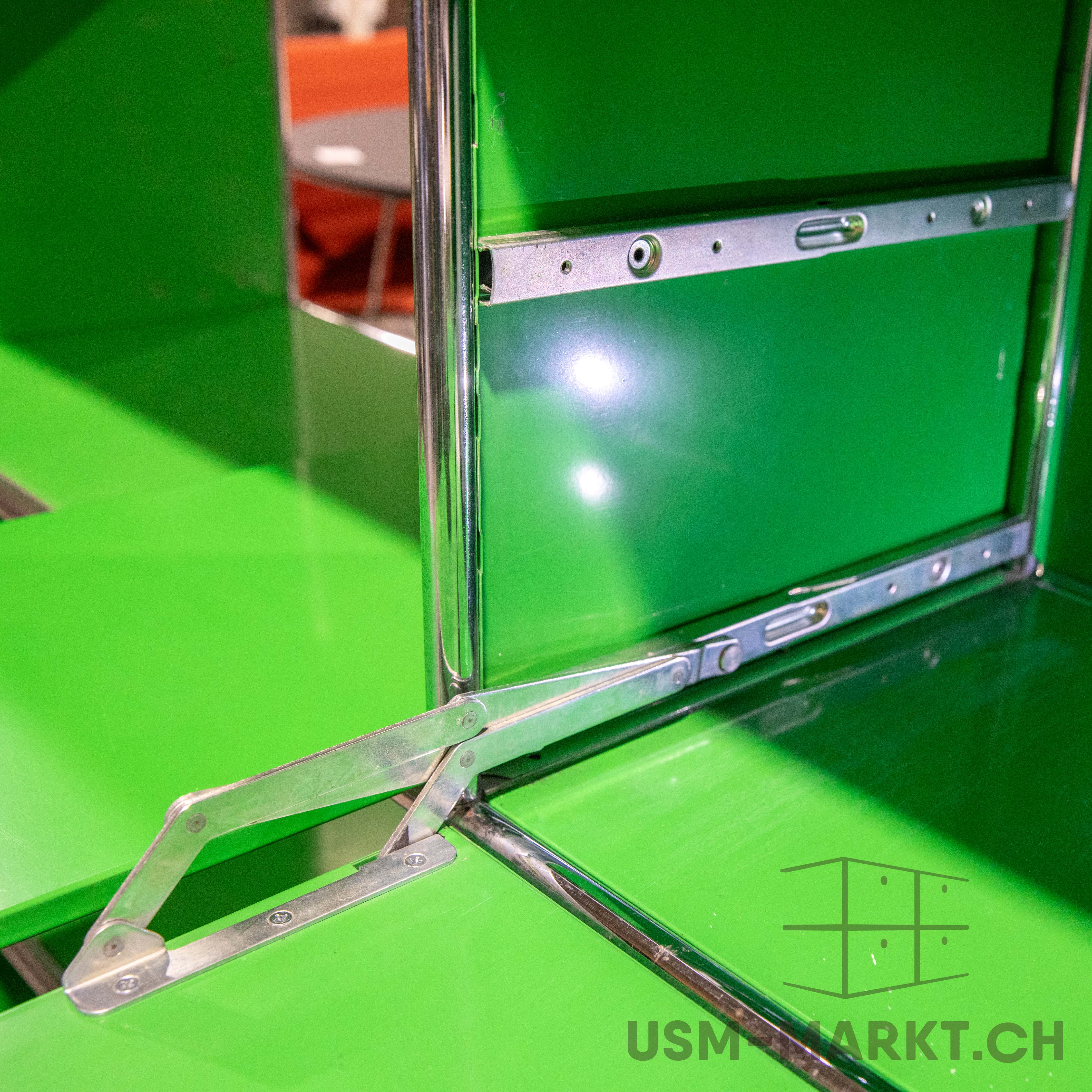 A close-up of a modular green USM Haller piece of furniture with polished chrome tubes and silver joints. An open joint connects two green panels at the front; in the background, another rail with fixings is visible.
