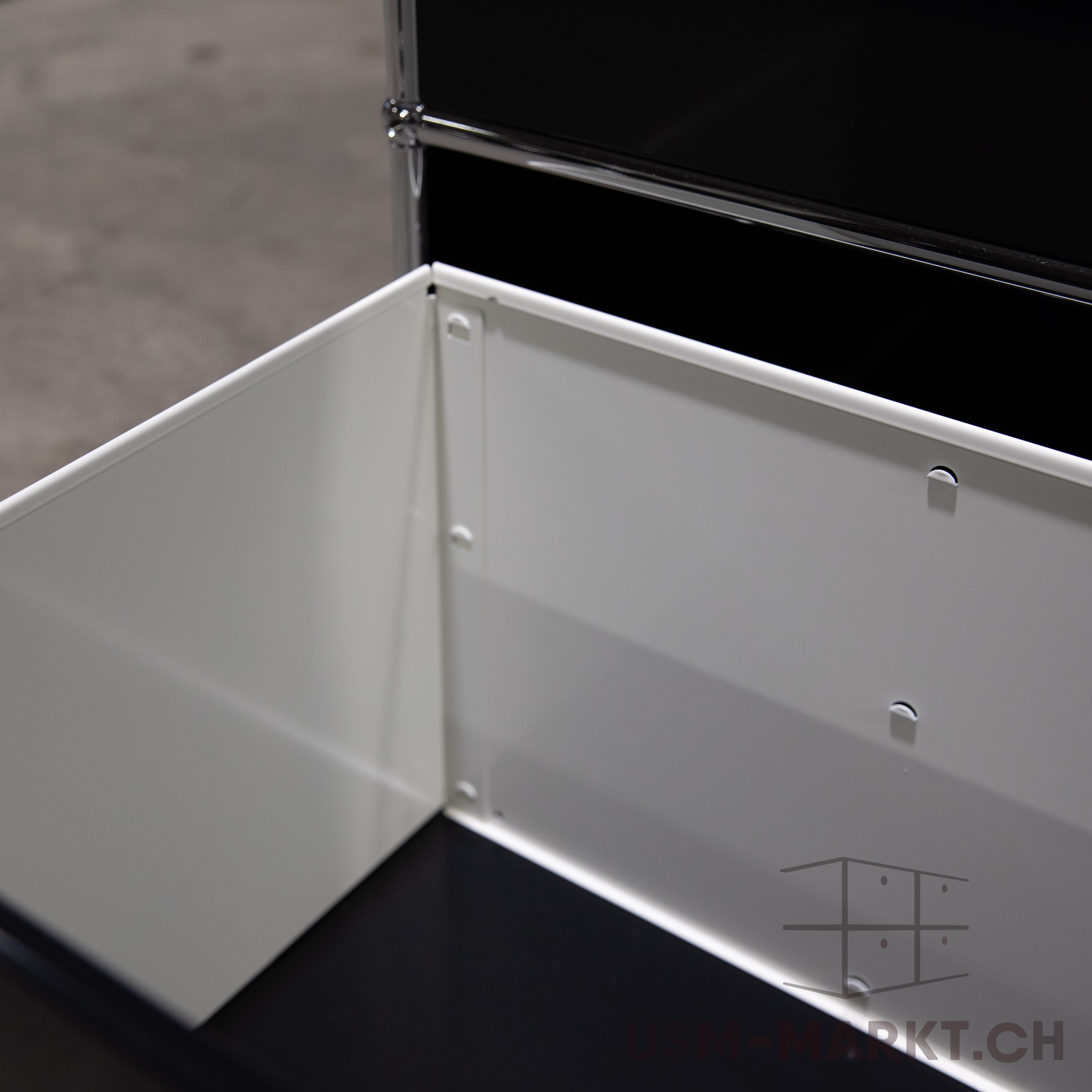 Nahaufnahme eines geöffneten, modularen Möbelstücks mit glänzenden weißen Innenwänden und dunklem Boden. An den Seitenwänden sind kleine runde Aussparungen für Befestigungspunkte sichtbar. Oben glänzt eine Chromstange am Rahmen.