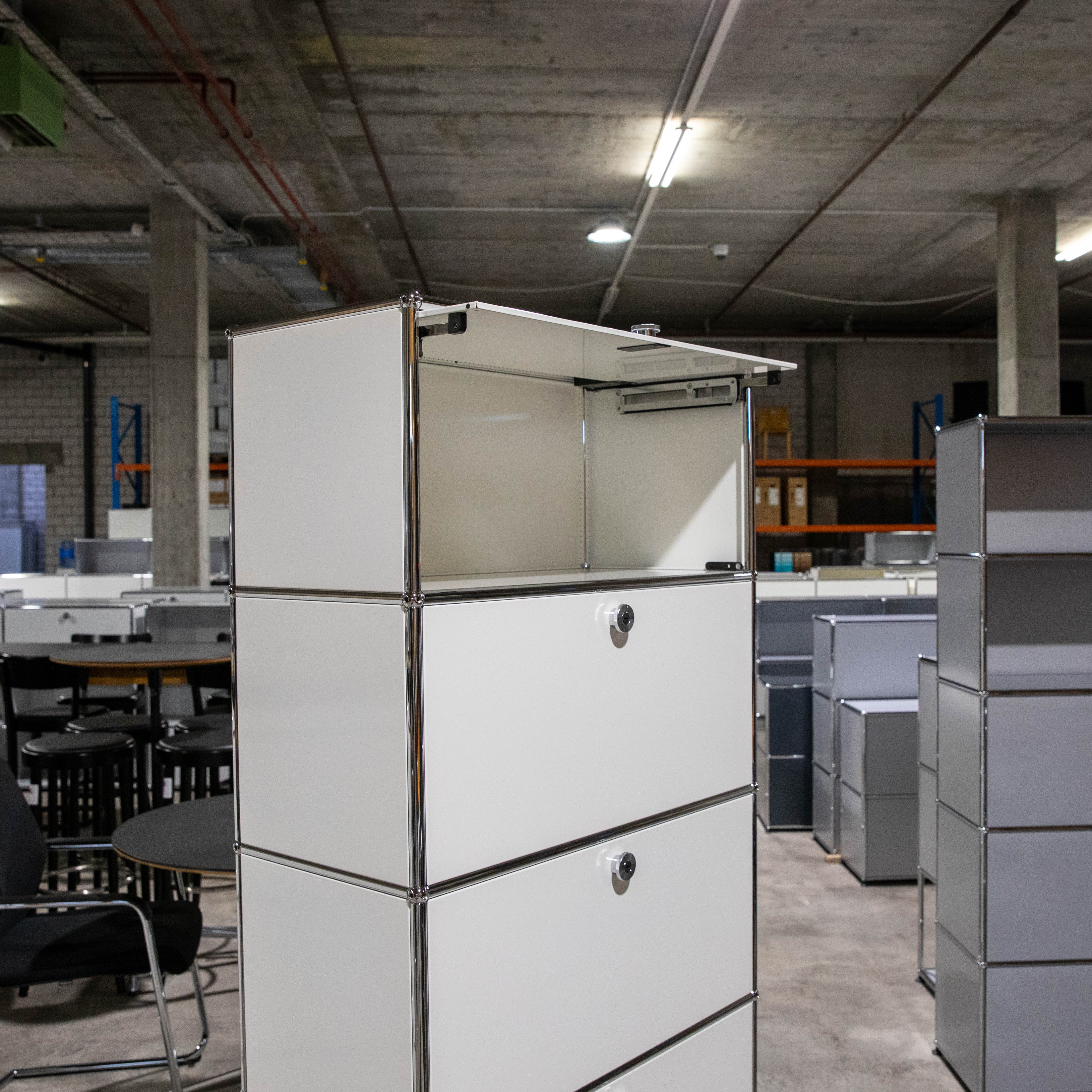 Weisses USM Haller Regal mit Chromrahmen. Oben offenes Klappfach, darunter drei geschlossene Fächer (teils mit Schloss). In Lager/Büro mit weiteren Möbeln, Betondecke und Leuchten.