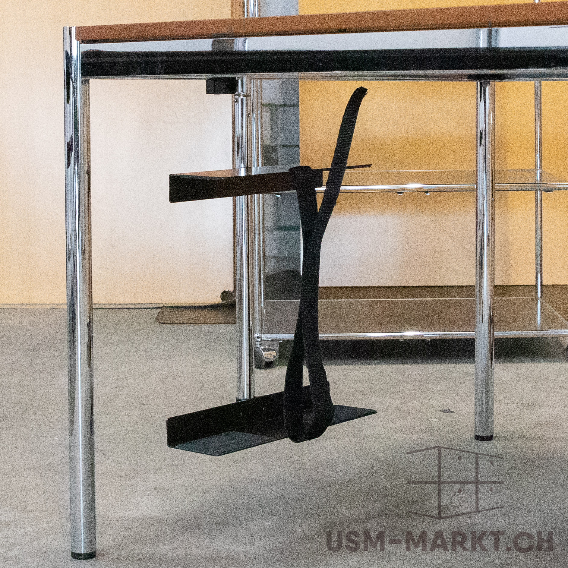 A detail of the underside of a table with polished chrome legs and struts supporting a wooden top. A black footrest hangs beneath it: a metal plate and a black fabric strap forming a loop, ready for use.