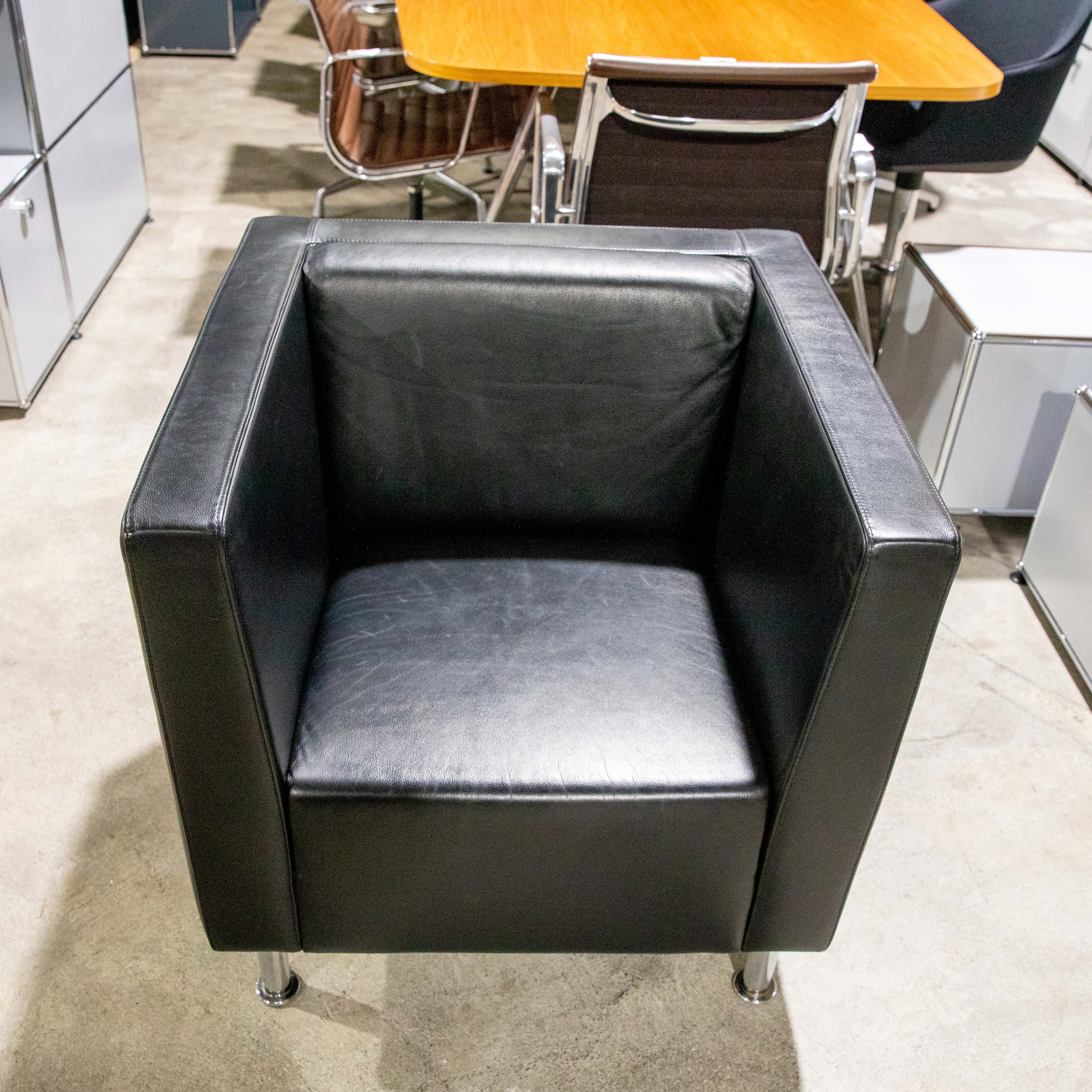 A modern black leather armchair positioned in the centre of a light grey concrete floor in an office. Cubic shape, thick upholstery, straight armrests. Short cylindrical legs in chrome. Out of focus: a wooden table and chairs.
