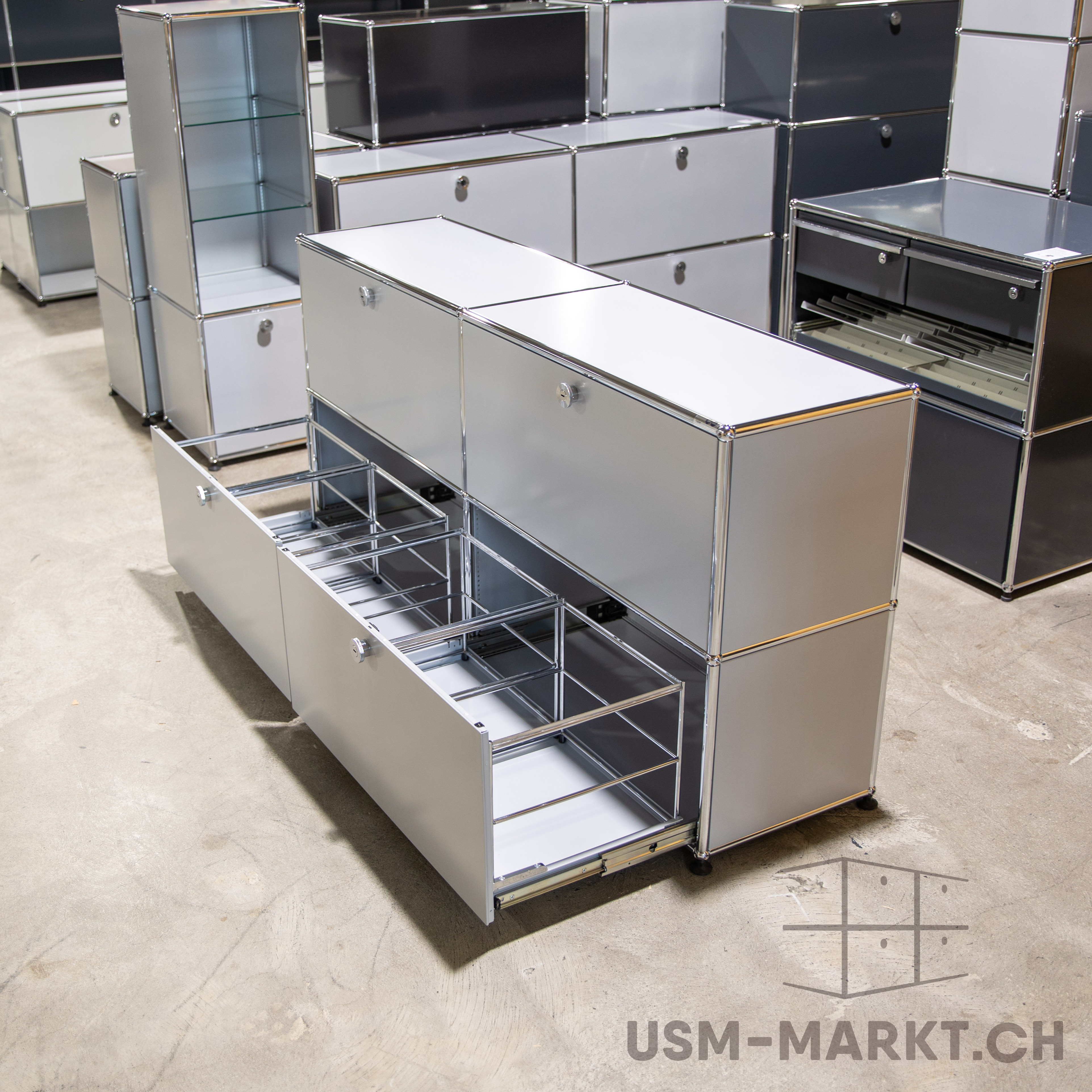 Modularer Schrank in hellem Grau mit Chromrahmen, unten drei Schubladen teils geöffnet, Metallauszüge sichtbar. Im Hintergrund weitere Module in Grau, Weiß und Schwarz, darunter ein hohes Regal mit Glasböden, auf Betonboden.