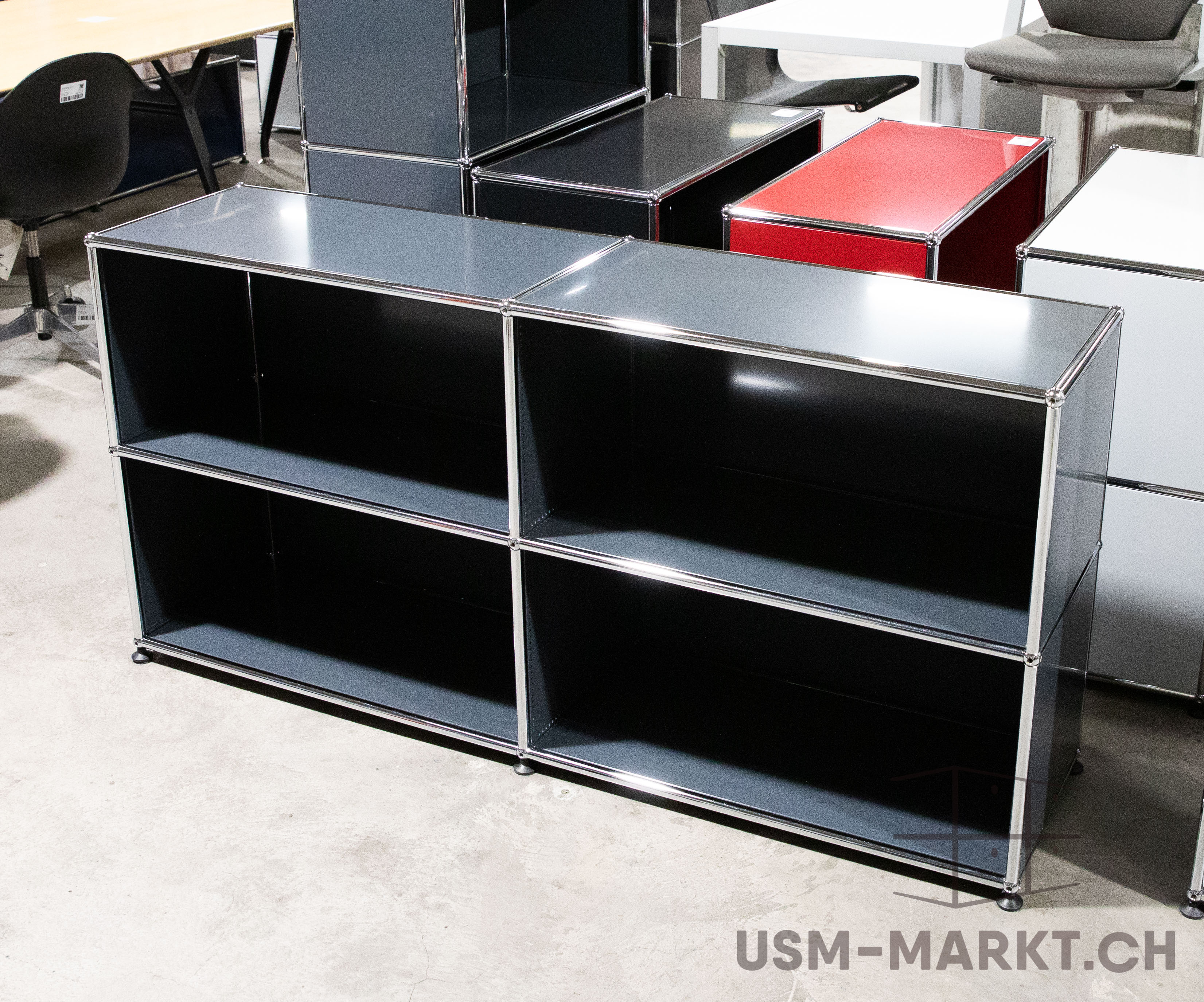 Schwarzes modulares Regal mit glänzendem Chromrahmen auf Betonboden. Zwei Reihen und zwei Spalten bilden vier offene Fächer. Im Hintergrund moderne Büromöbel sowie Regalelemente in Rot und Weiß, wie in Ausstellung oder Geschäft.