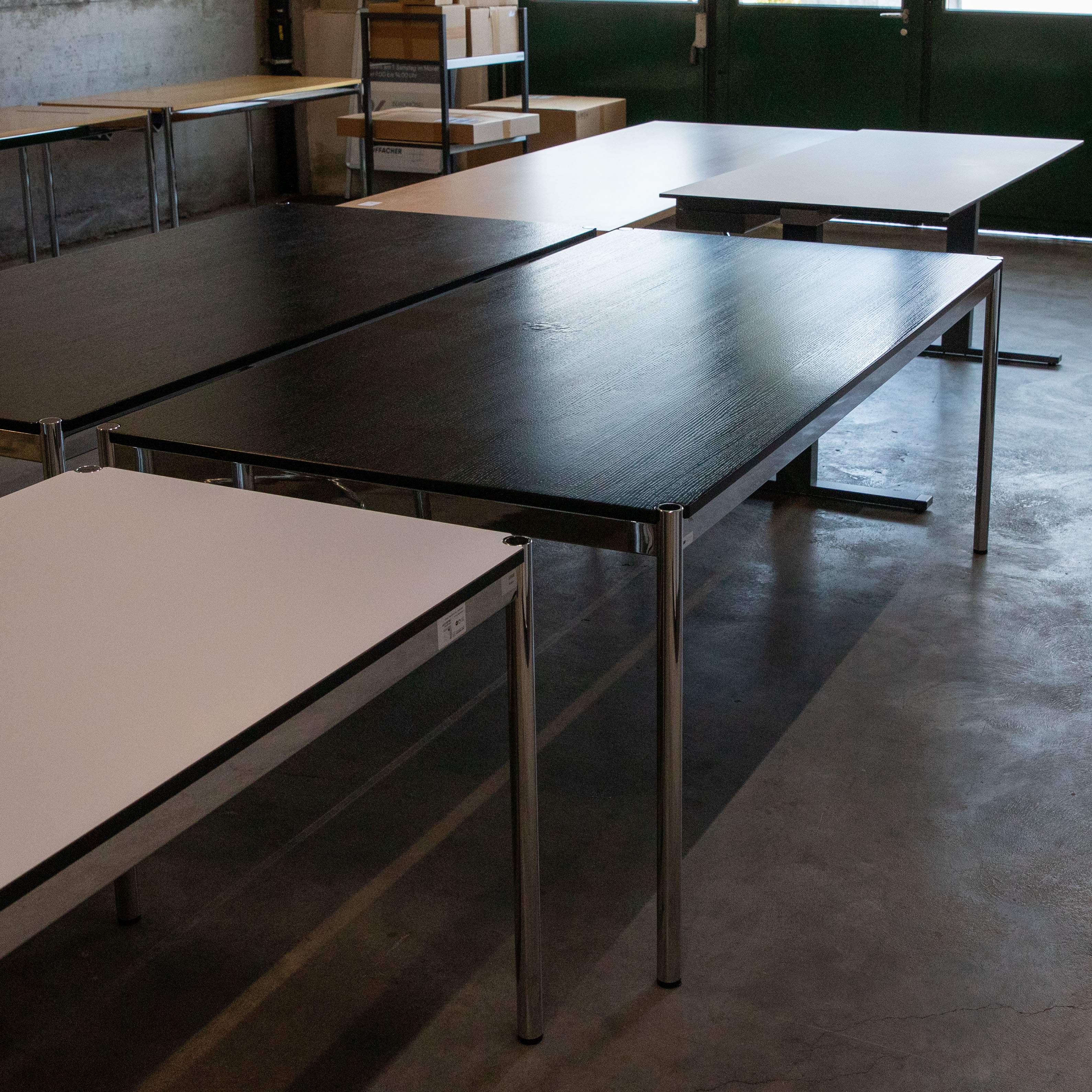 Second-hand USM Haller table (200 x 100 cm) with a black oak top and polished chrome legs on a concrete base. The top shows clear signs of water damage: several raised blisters. Other tables can be seen in the background.