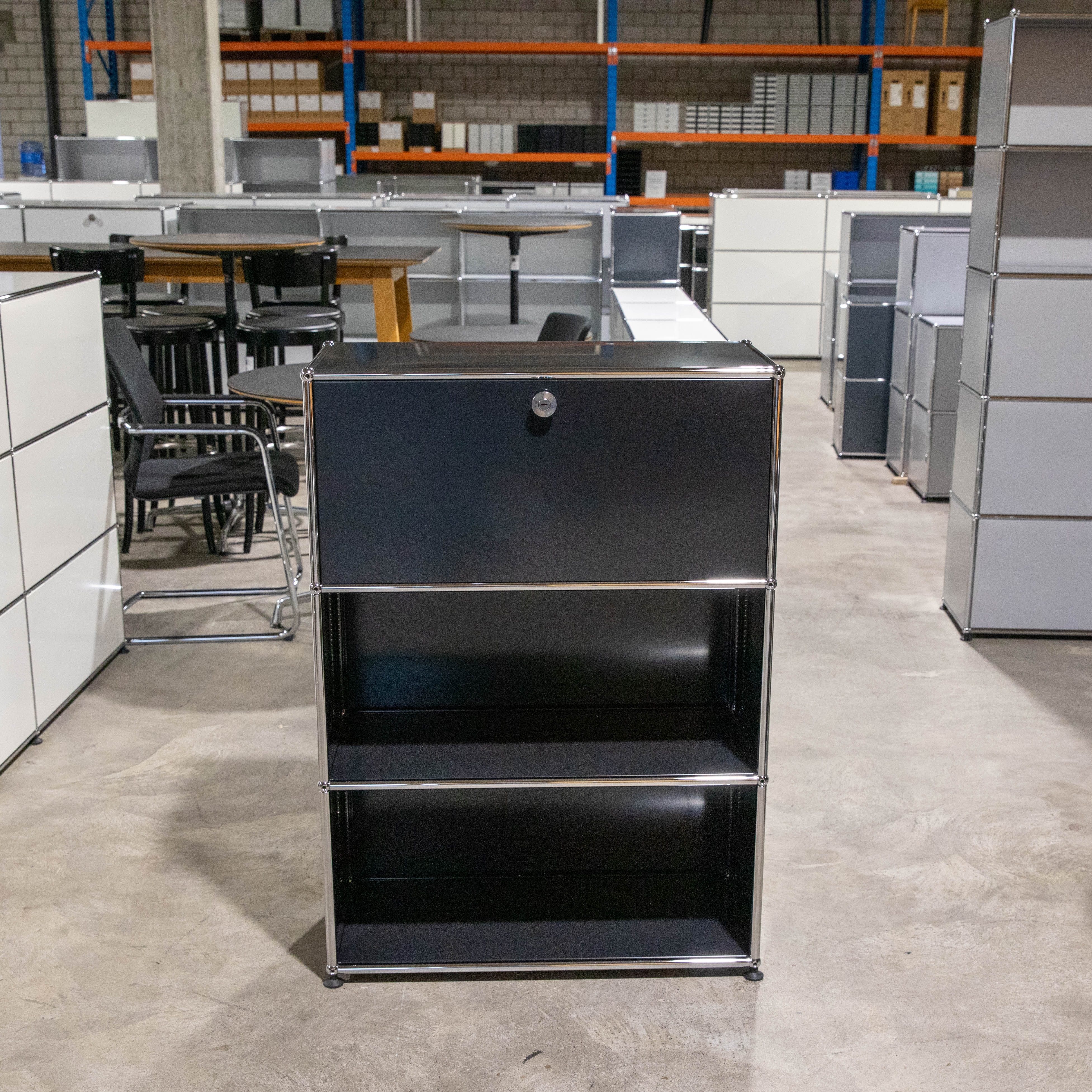 Schwarzes USM Haller Regal mit drei Ebenen auf Betonboden. Oben geschlossene Klapptür mit Schloss, darunter zwei offene Fächer. Modulares Möbel mit glänzendem Chromgestell. Im Hintergrund weitere helle USM Haller Möbel und Lagerregale.