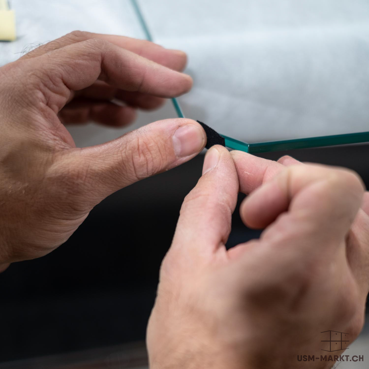 Due mani fissano una piccola striscia di feltro nero o di gomma all'angolo di un pannello di vetro trasparente. Il bordo riflette una luce verdastra e mette in evidenza lo spessore del materiale. L'attenzione è concentrata sull'azione e sul materiale.