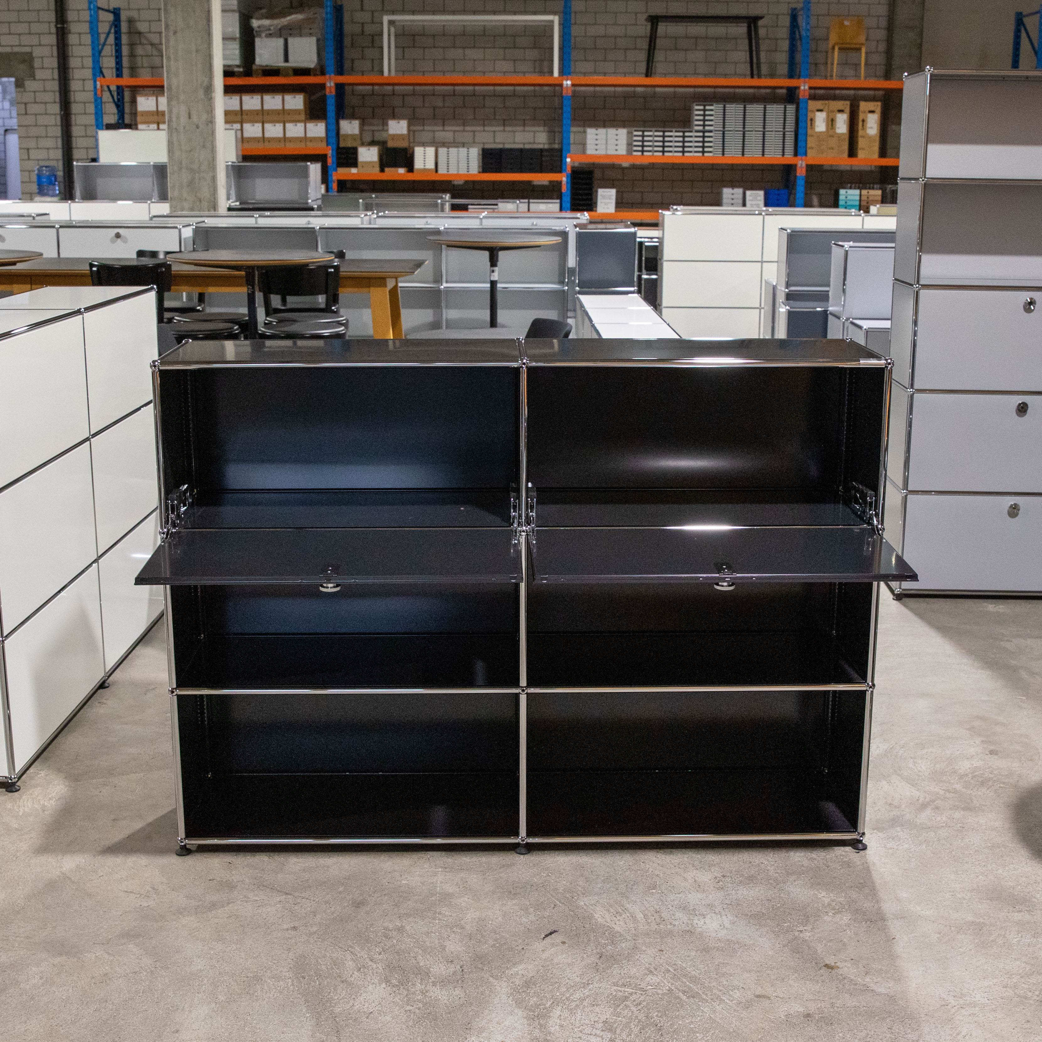 Schwarzes USM Haller Regal mit drei Reihen und zwei Spalten. Die oberen beiden Fächer sind als Klapptüren geöffnet und zeigen den leeren Innenraum. Das modulare Möbel steht auf Betonboden im Lager, umgeben von Regalen.