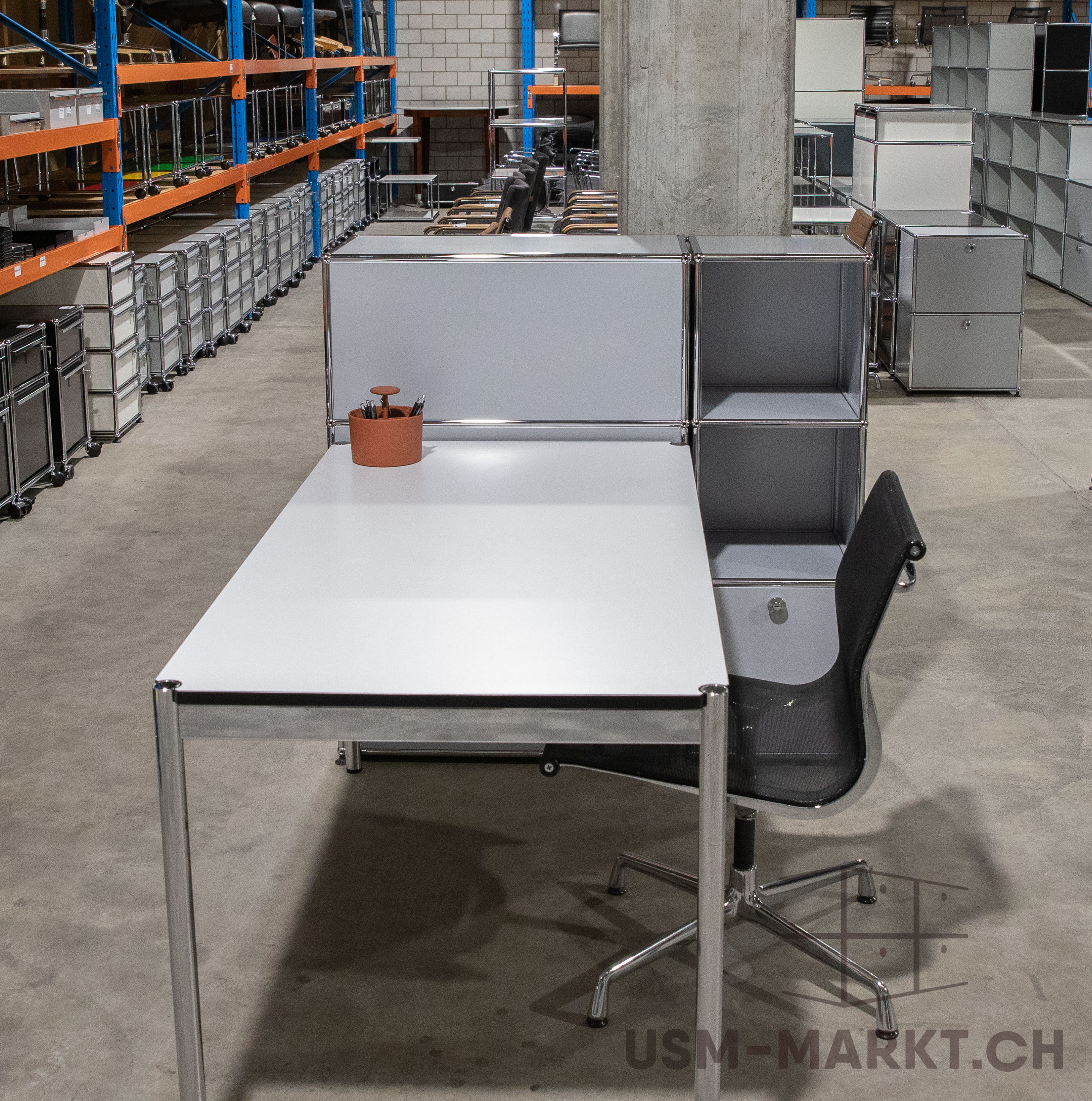 Modernes weißes USM Haller Schreibtischsystem mit schwarzem Bürostuhl in großem Lagerraum. Dahinter modulare USM Haller Regalelemente in Hellgrau. Im Hintergrund weitere USM Haller Möbel und Rollcontainer an hohen orangen/blauen Regalen.