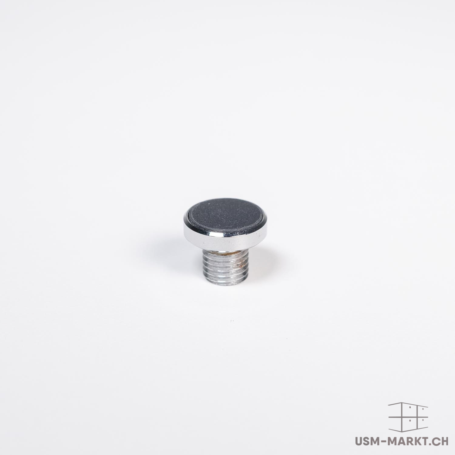 A small, shiny metal foot or spacer with a threaded base and a round, matt black or dark grey disc on top, edged by a narrow, silver ring. Set against a neutral white background.