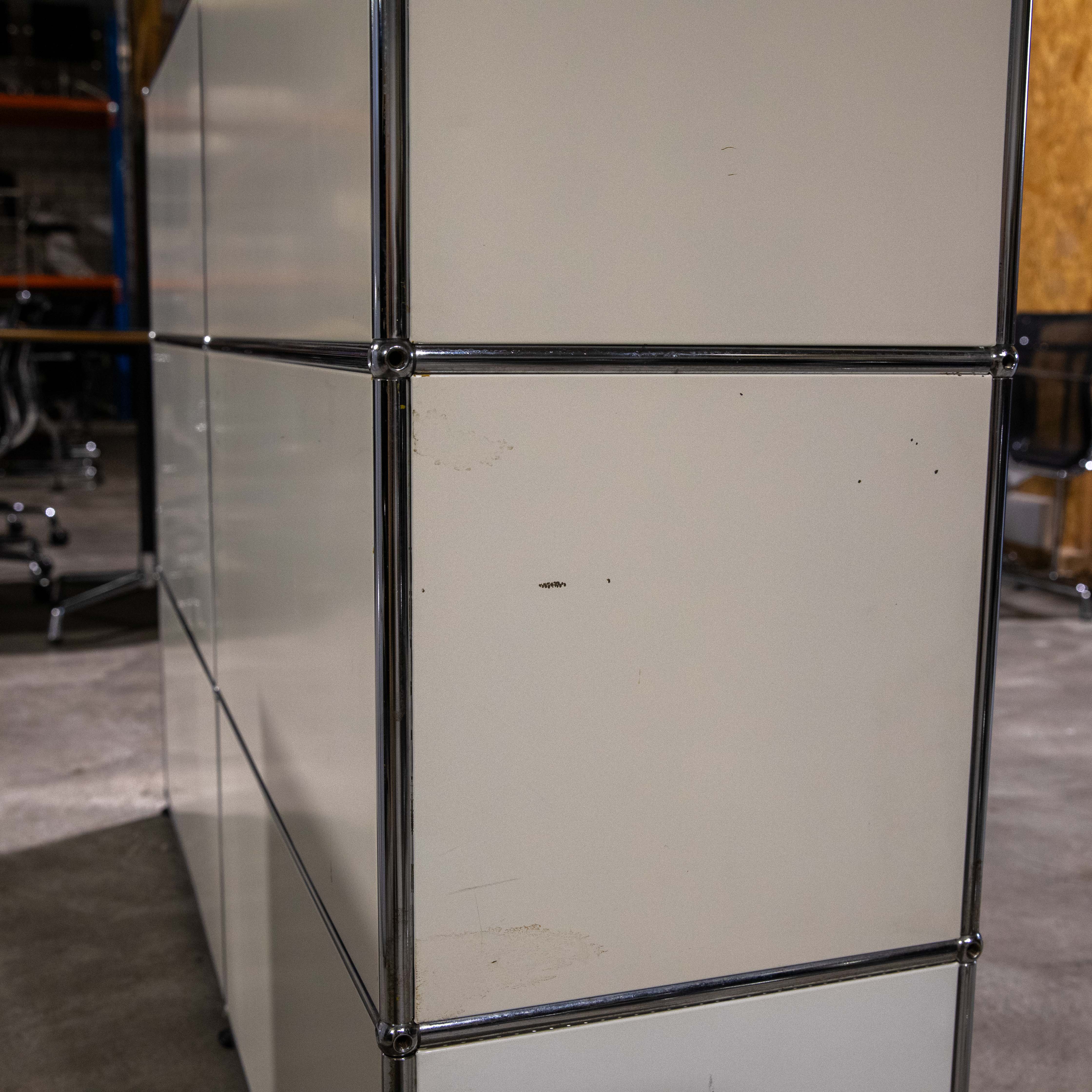 Modulares Regal aus cremefarbenen Metallpaneelen und glänzenden Chromstahlrohren in einem Innenraum. Symmetrisch angeordnete Fächer mit leichten Gebrauchsspuren wie kleinen Flecken. Im unscharfen Hintergrund weitere Möbel.