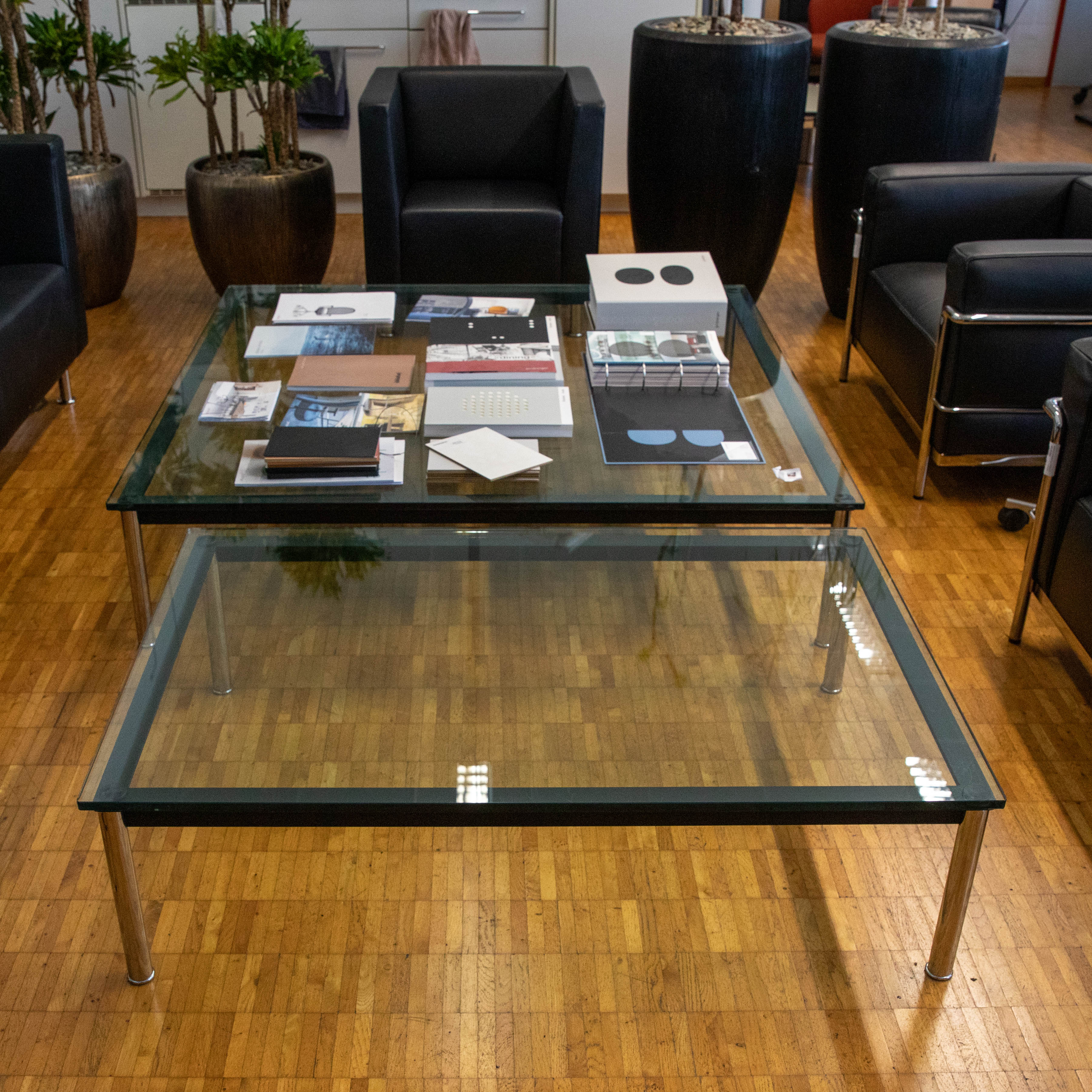 Deux tables rectangulaires en verre dans une pièce moderne dotée d'un parquet en bois. Sur la table du haut se trouvent des brochures, des papiers et des carnets. À côté, on trouve des fauteuils en cuir noir et de grandes plantes dans des pots foncés.